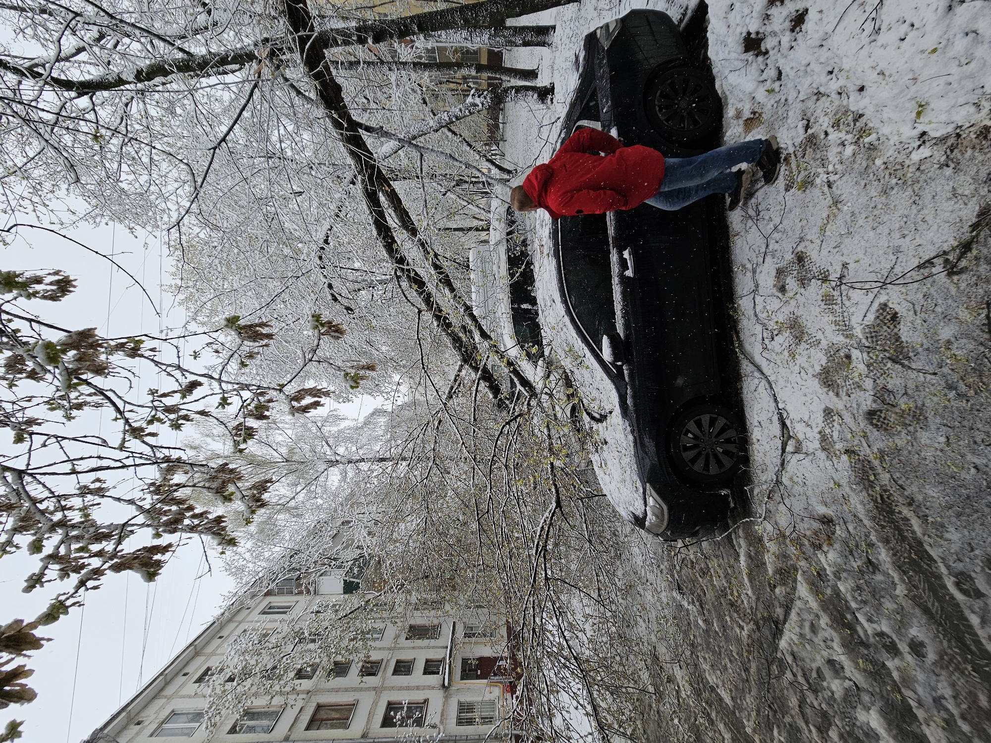 A person in a red jacket and blue jeans stands on a snowy street in Khimki, Russia, next to a black car. The person is leaning over the car's hood. The street and surrounding areas, including trees and buildings, are covered in ice and snow. A multi-story building with numerous windows is visible in the background.