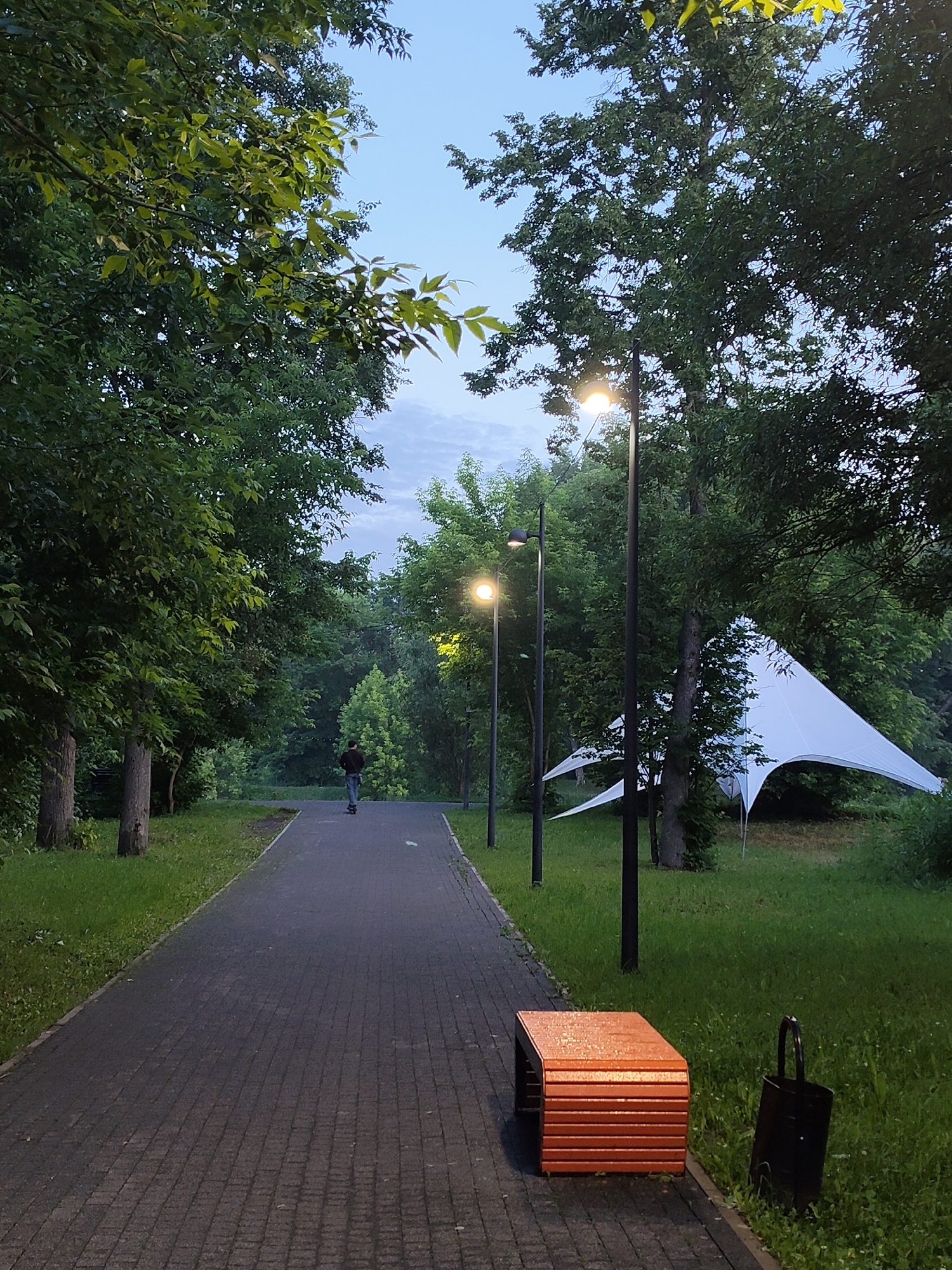 An outdoor scene at dusk or dawn depicts a paved pathway lined with grass and trees. A person is riding an electric scooter on the pathway in the distance. Streetlights are visible along the path, illuminating the surroundings. To the right of the pathway, a white tent structure is set up amongst the trees. In the foreground, an orange bench and a black trash can are placed on the grass. The sky is overcast with hints of blue. The location is described as being in the city of Chekhov, Russia.
