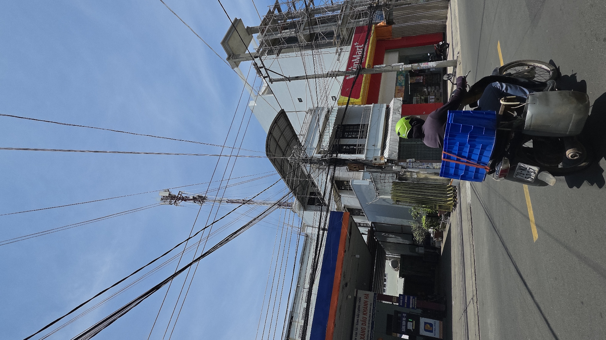 The image shows a street scene in Nha Trang, Vietnam. A man on a motorbike wearing a lime-green helmet is facing away from the camera and riding to the right of the frame along a paved road. The bike has a blue plastic crate tied to the back. The buildings on the left feature tangled power lines, a VinMart store, and a petrol station ("Xăng dầu Khánh Hà"). The sky is clear and blue.