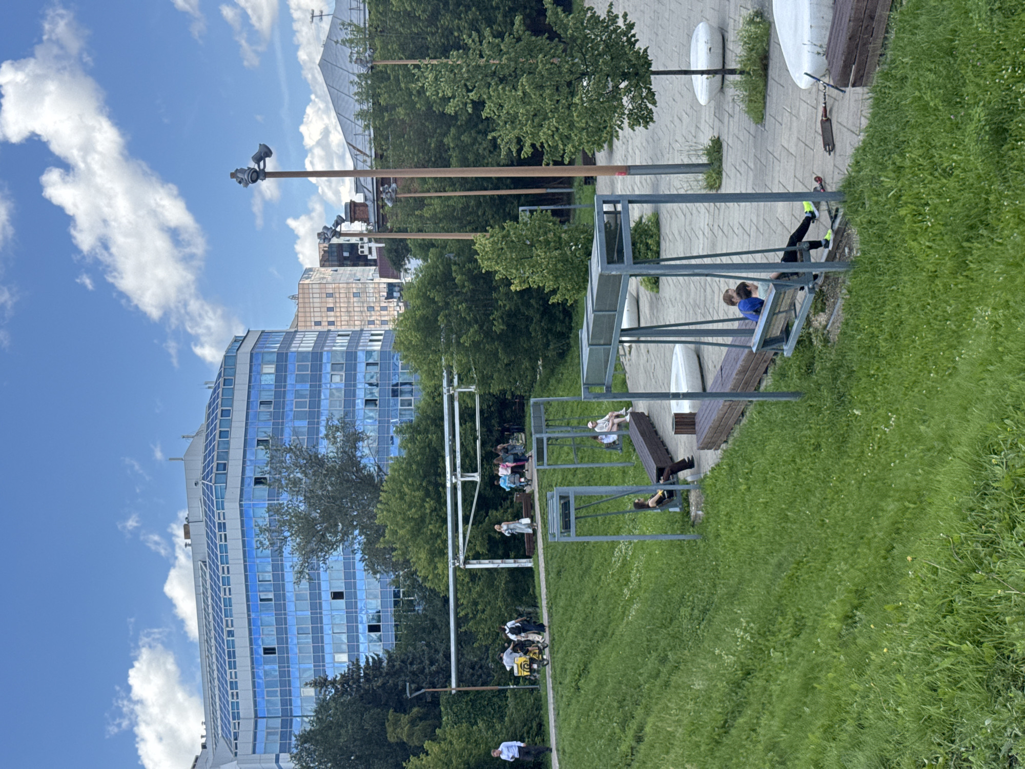 The image depicts a public park area with modern architecture in the background. A multi-story building with a blue glass facade is prominent. The park itself features a grassy slope in the foreground, with a paved area and modern park amenities in the midground, including swings, benches, and stylized white seating structures. Several people are visible throughout the scene: one individual in a suit walks on a path, a group of people are near a motor scooter, and several individuals are seated on benches or swings. Some people appear to be resting or socializing. The environment is outdoors under a partly cloudy sky. This location is identified as a park in Yekaterinburg, Russia, specifically near the building "Krylova 27".