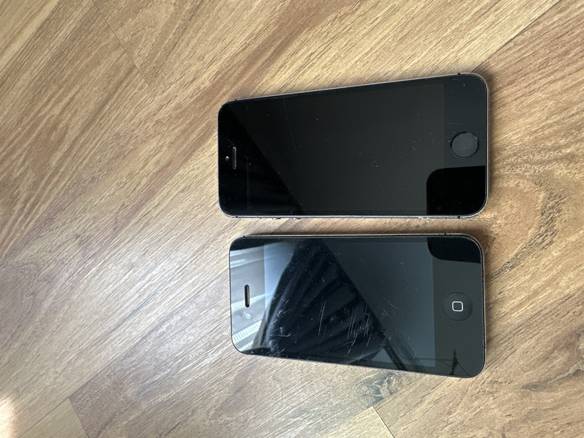 Two dark-colored smartphones are positioned side-by-side on a light brown wood-grain laminate floor in Unknown, Unknown. Both phones are oriented vertically with their screens facing upwards. The phone on the left, an iPhone 4 or 4S, features a black front panel with a home button marked by a white square icon. Its screen displays numerous prominent scratches. The phone on the right, an iPhone 5-series or SE (1st generation), also has a black front panel but features a circular, unadorned home button. Its screen has fewer and less prominent scratches compared to the phone on the left. Both devices exhibit visible earpiece cutouts and front-facing cameras near their top edges. The laminate floorboards run diagonally from the lower-left to the upper-right of the frame. The lighting suggests ambient light, creating reflections on the phones' screens and the floor surface. No individuals or other objects are present within the frame.