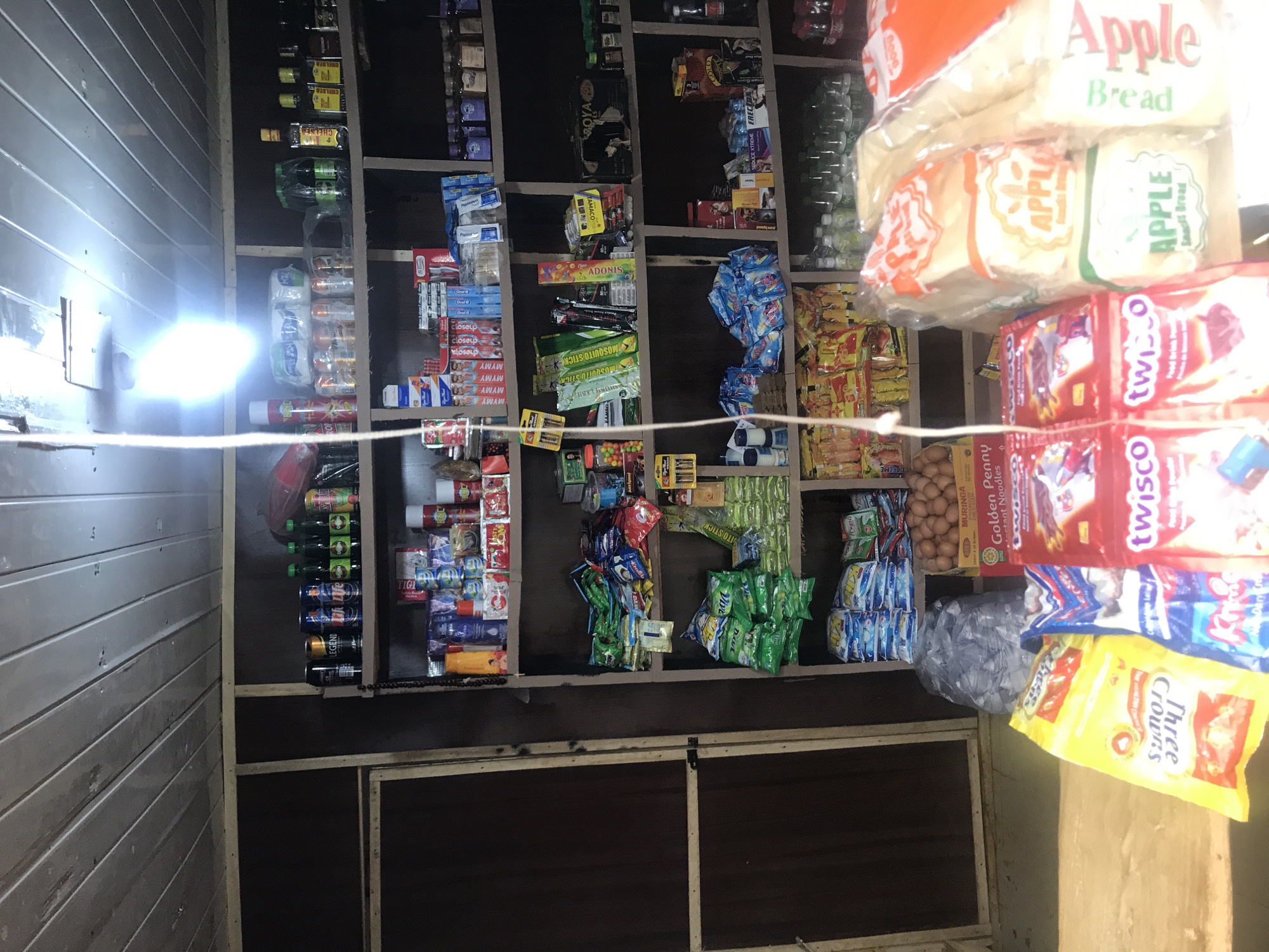 The image depicts a small, dimly lit convenience store or local market stall, likely in New Stadium, Nigeria. The shelves are packed with a variety of goods, including packaged foods, drinks, and household items. On the top shelves, bottles of alcoholic beverages and soft drinks are visible, alongside some canned goods. The middle shelves are filled with smaller packaged items such as snacks, toiletries, and cleaning supplies. The bottom shelves and the foreground display a mix of packaged foods, including bread, biscuits, and what appear to be instant noodles. A box of eggs is also visible on a lower shelf, along with a large plastic bag of what might be water sachets. The overall impression is of a well-stocked, albeit somewhat cluttered, local shop. There are no people visible in the image.