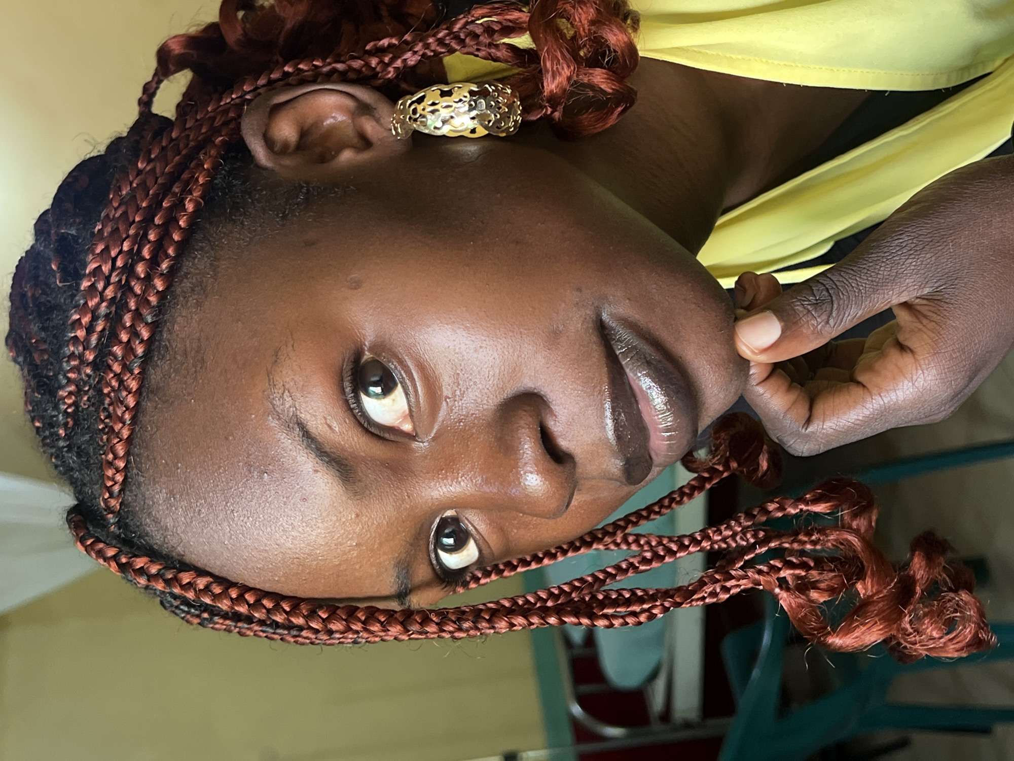 The image shows a close-up, sideways portrait of a young Black woman. She has braids that appear to be a reddish-brown color, with some strands twisted and others loose. She is wearing a bright yellow garment. A gold, patterned ring is visible on her right ear. Her skin is a rich, dark brown, and her lips are glossy. Her eyes are open and looking towards the camera, with a hint of a smile. One of her hands is visible, with fingers touching her chin. The background is softly blurred, suggesting an indoor setting with muted colors. The lighting appears to be natural, creating soft shadows on her face. No text is visible in the image. The overall mood is one of warmth and gentle curiosity.