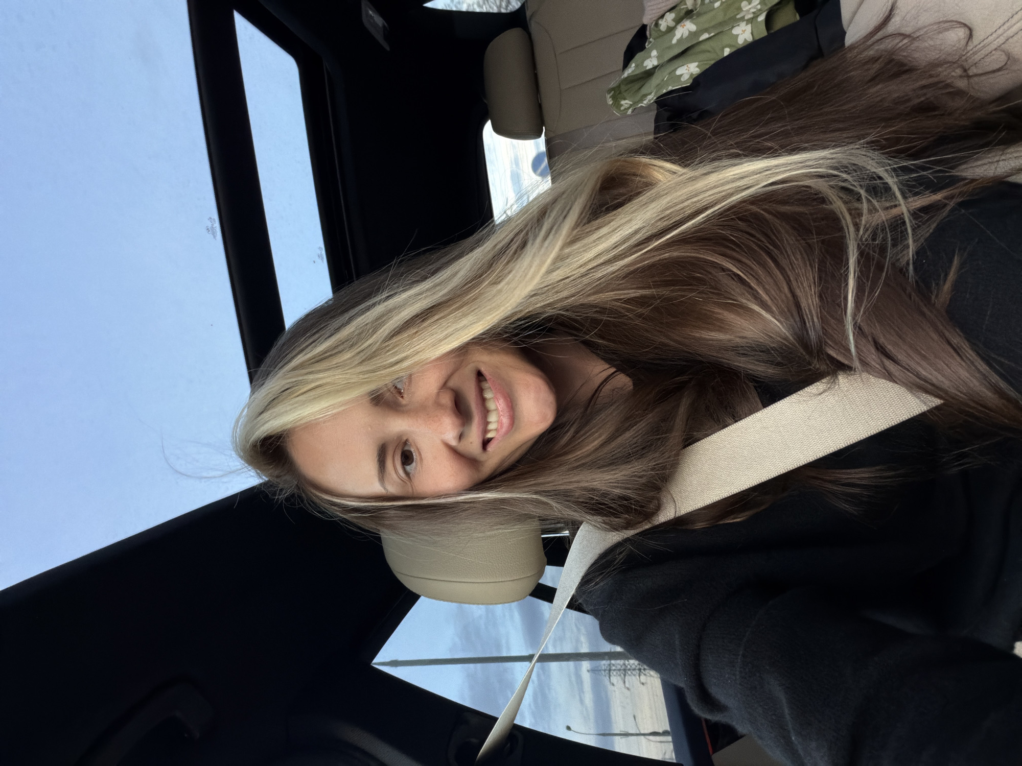 A young woman with long, highlighted hair is wearing a seatbelt in a car, smiling at the camera. The car has a sunroof. The location appears to be outside of a city with what looks like a highway or bridge visible through the car window. This scene does not appear to be in Moscow, Russia as there are no recognizable landmarks or architectural details consistent with that location. The environment suggests a more open or suburban setting, possibly near a highway or rural area.