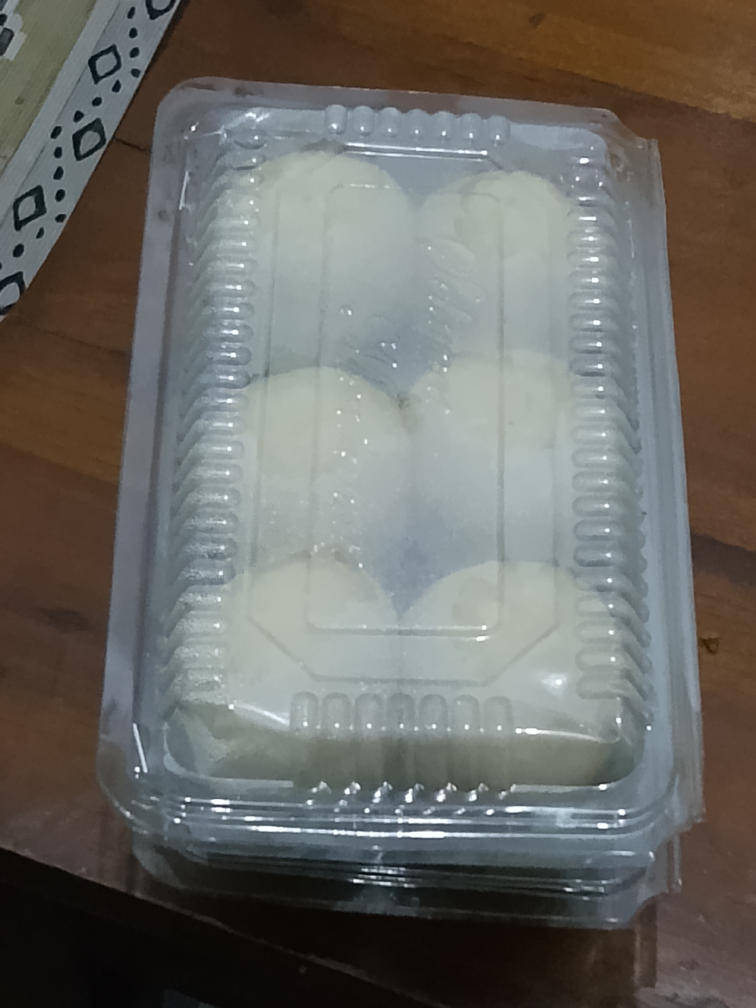 A clear plastic container holding six white, round pastries is placed on a wooden surface. The container is rectangular with ribbed sides and a clear lid. The pastries appear to be dusted with a light powder. In the upper left corner of the image, a portion of a patterned object with black diamond shapes and dots on a light background is visible. The scene is likely indoors, on a table, in Betonan, Indonesia.