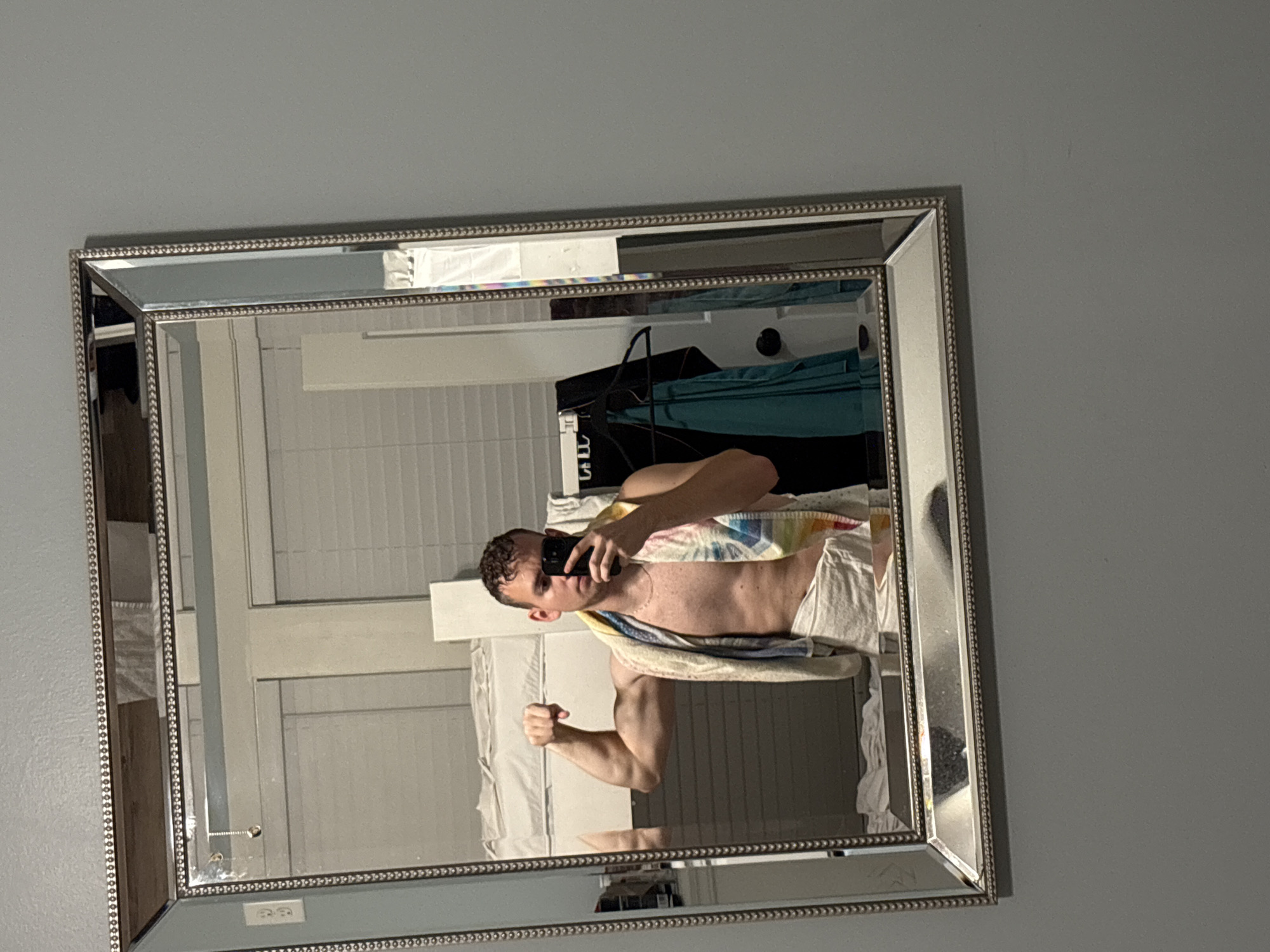 A young adult male is standing in front of a mirror, taking a selfie. He is flexing his bicep and wearing a towel around his waist. A colorful towel is draped over his shoulders. The mirror reflects a room with bunk beds and clothing hanging on a rack. The image is taken indoors in what appears to be a bedroom or dorm room. The walls are painted a neutral gray color. The mirror has a decorative silver frame. The location is Panama City Beach, United States of America.