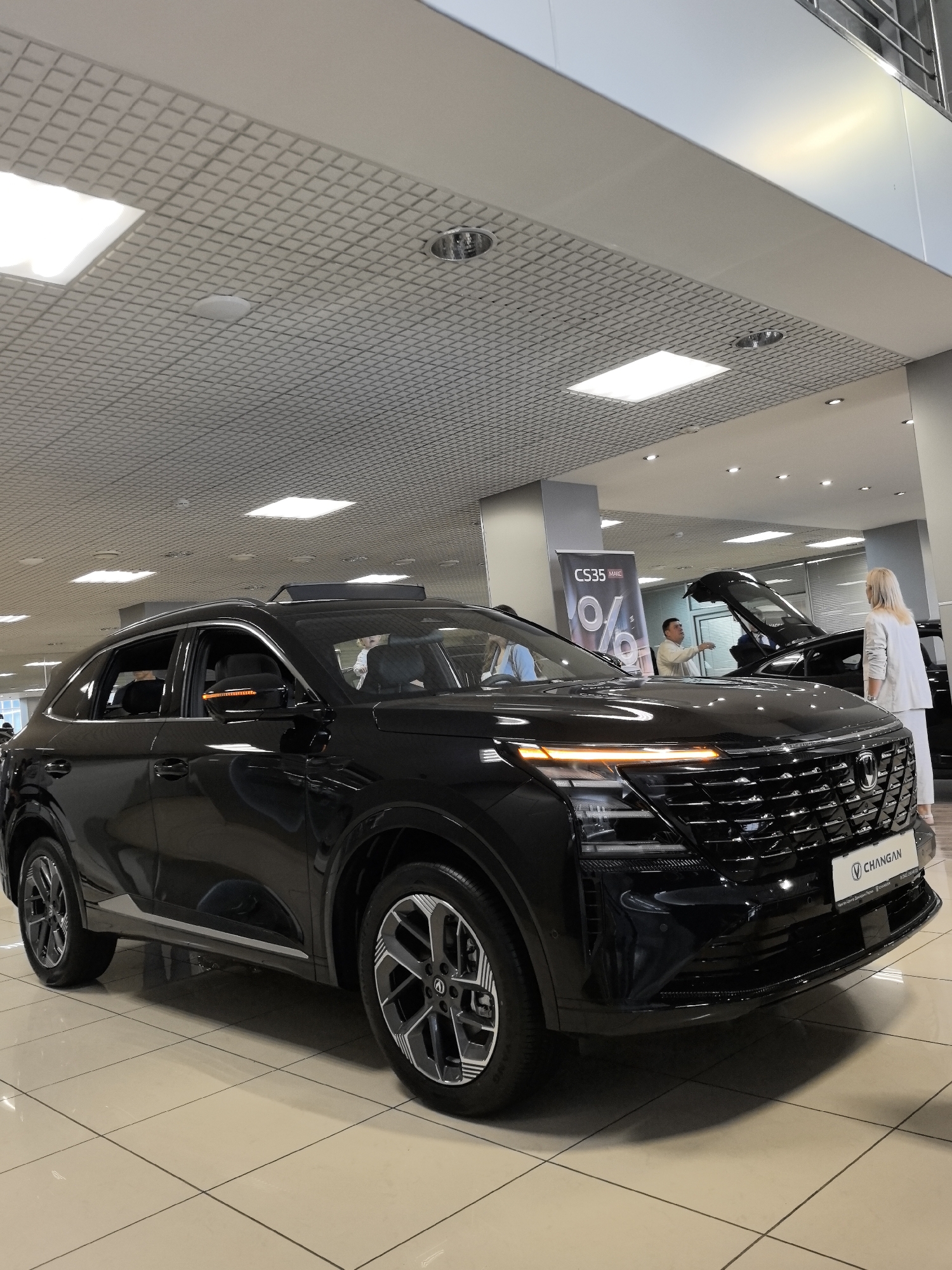 The image depicts a black Changan CS35 PLUS automobile on display inside a brightly lit car dealership. The car's front fascia, grille, headlights, and wheels are prominently visible. The dealership's interior features a tiled floor, a suspended ceiling with integrated lighting, and several large windows. In the background, a banner advertising "CS35 MAX" with percentage signs is displayed. Two individuals, a man and a woman, are present in the scene; the man appears to be gesturing towards another vehicle, possibly discussing it with the woman who is facing away from the camera. The overall environment suggests a commercial setting, specifically a car showroom. The location is confirmed to be Perm, Russia.