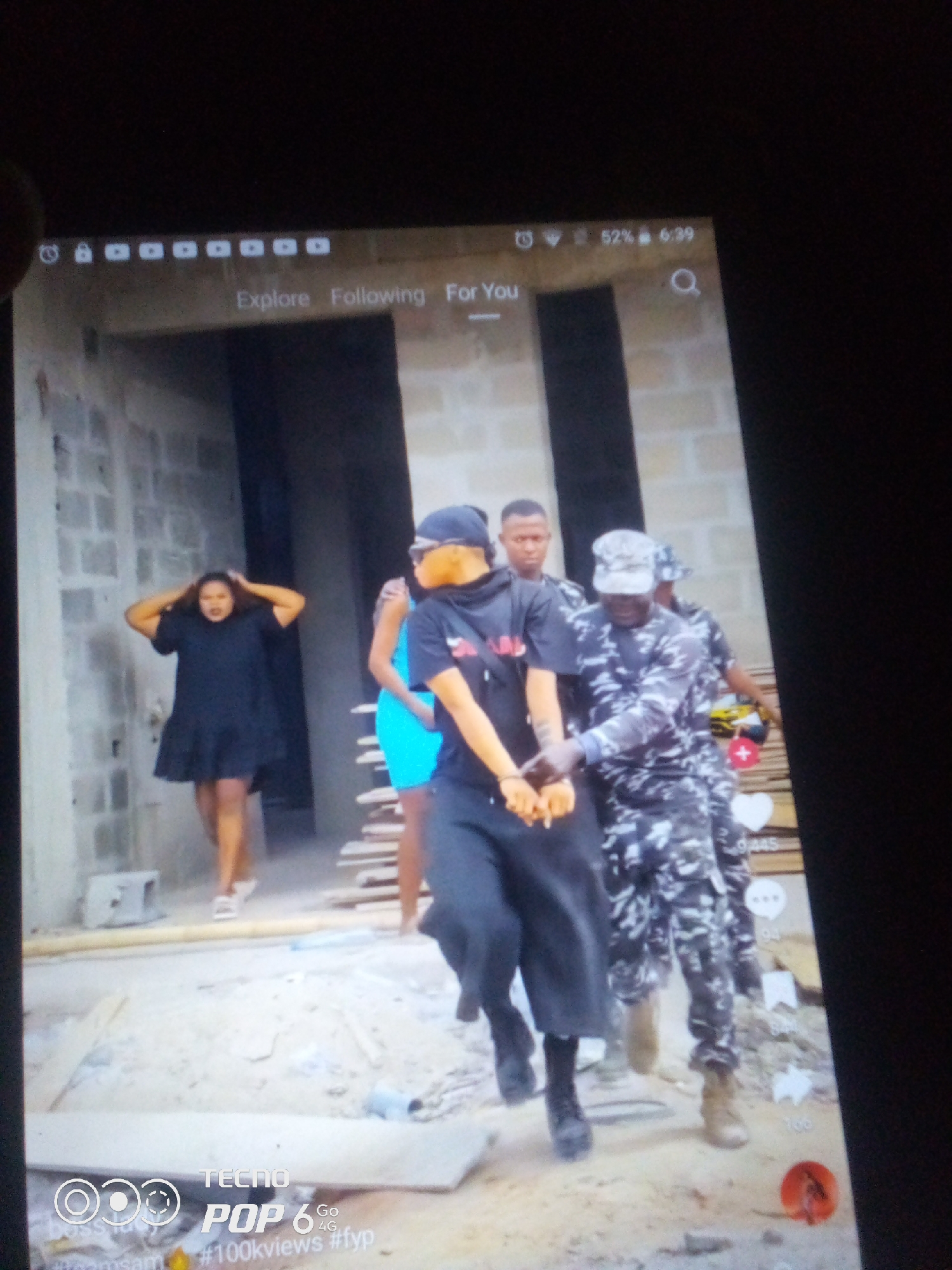 The image, captured on a mobile device, depicts an outdoor scene with several individuals, possibly in a construction or unfinished building site. In the foreground, a woman in a black dress is seen with her hands on her head, looking distressed. She is accompanied by at least three other people, two of whom are wearing camouflage-patterned uniforms, suggesting they might be law enforcement or security personnel. One of these individuals is holding the woman's hands, while another stands behind him. The environment appears to be dusty and unkempt, with building materials and debris visible. The upper portion of the image shows a mobile device interface with icons and text, including "Explore," "Following," "For You," and what appears to be network and battery status. The location of Howayi, Nigeria, is not discernible from the visual elements within the image itself.
