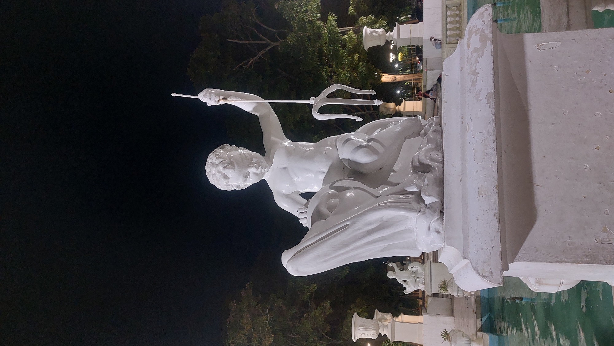 The image shows a white statue of Neptune holding a trident, mounted on a pedestal within a fountain. The statue is located in Plaza Venezuela, Caracas, Venezuela. Other people are standing around the fountain in the background.