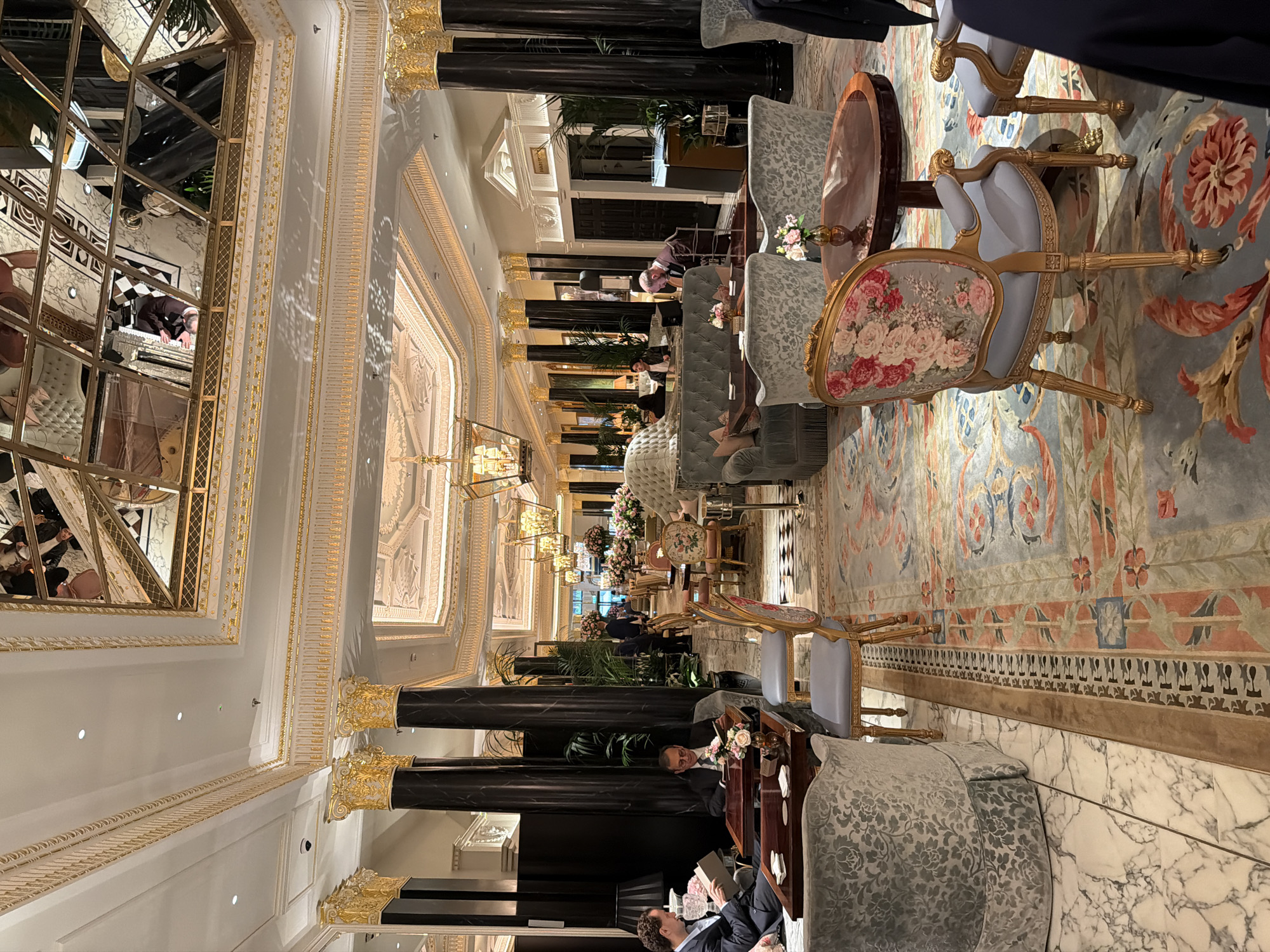 The image captures the opulent interior of a high-end establishment, most likely a hotel or a restaurant in London, United Kingdom. The scene presents a long, richly decorated hallway with ornate ceiling moldings and decorative pillars, suggestive of a classic and luxurious setting.
In the foreground and throughout the hallway, there is arranged seating, including upholstered chairs and sofas with accompanying small tables. Several individuals are present, some seated and engaged in conversation or reading, others are standing or walking in the distance.
The color palette is dominated by golds, blues, and soft neutral tones, contributing to the overall impression of elegance and sophistication. The floor is partially covered by an intricate rug, and the lighting is warm and inviting, enhancing the atmosphere.