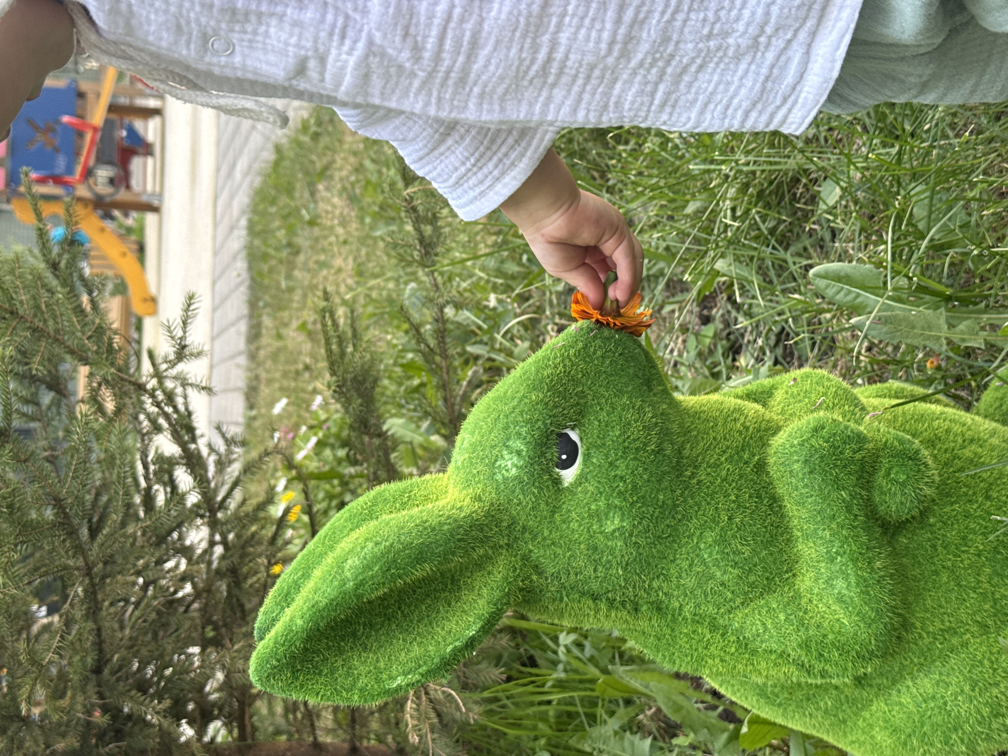 The image depicts an outdoor scene in an urban green space, characteristic of a park or residential courtyard in Moscow, Russia. In the foreground and midground, a green topiary rabbit, made of artificial grass-like material, is positioned on the left, facing right. It has a single visible black eye. To the right, a partially visible child, wearing a white textured long-sleeved shirt and light green trousers, extends their right hand towards the rabbit. The child is holding a small orange marigold-like flower between their thumb and forefinger, presenting it to the rabbit's nose area. The subjects are situated on a patch of green grass with some small weeds. Behind the rabbit, a coniferous tree with dark green needles is visible. In the background, beyond a paved surface, a children's playground with colorful equipment, including a yellow slide and blue and red structures, is present.