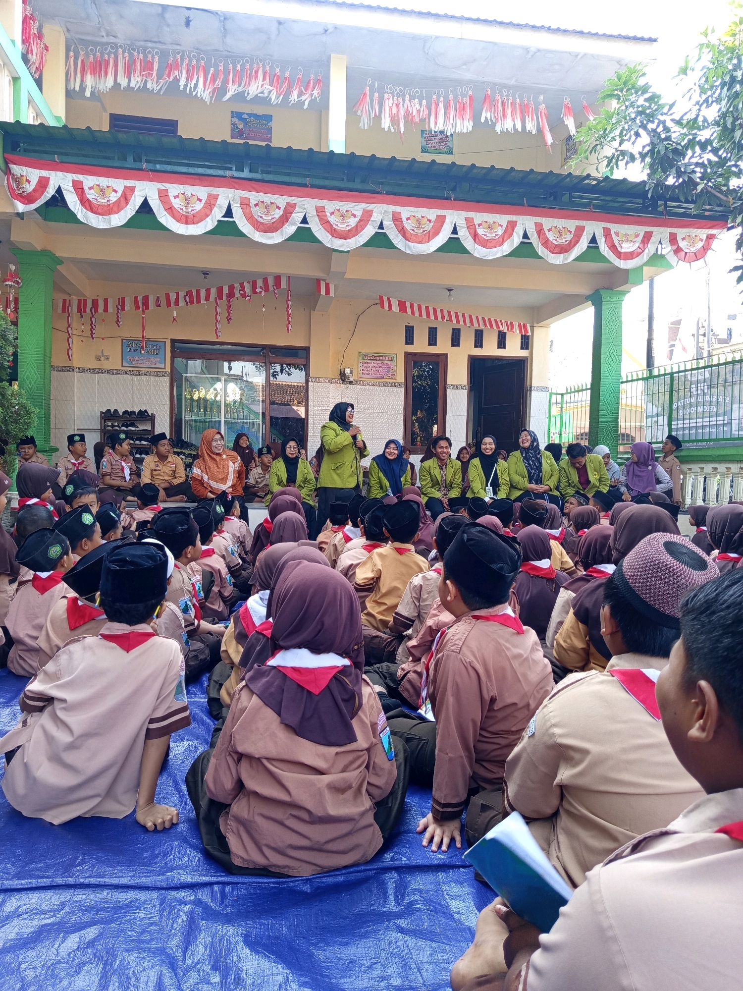 The image depicts an outdoor gathering, likely a school event, in Randubango, Indonesia. A group of children and adults are seated on a blue tarp spread on the ground, facing a raised platform or stage area. The children are dressed in scout uniforms, consisting of brown shirts and pants, with some wearing red and white neckerchiefs. The adults, some of whom are also in uniform-like attire, are seated or standing. One woman in a green jacket is speaking into a microphone. The background shows a building decorated with Indonesian flags and red and white banners, suggesting a national holiday or celebration. The environment is sunny, with trees visible to the right. The overall scene conveys a sense of community and organized activity.