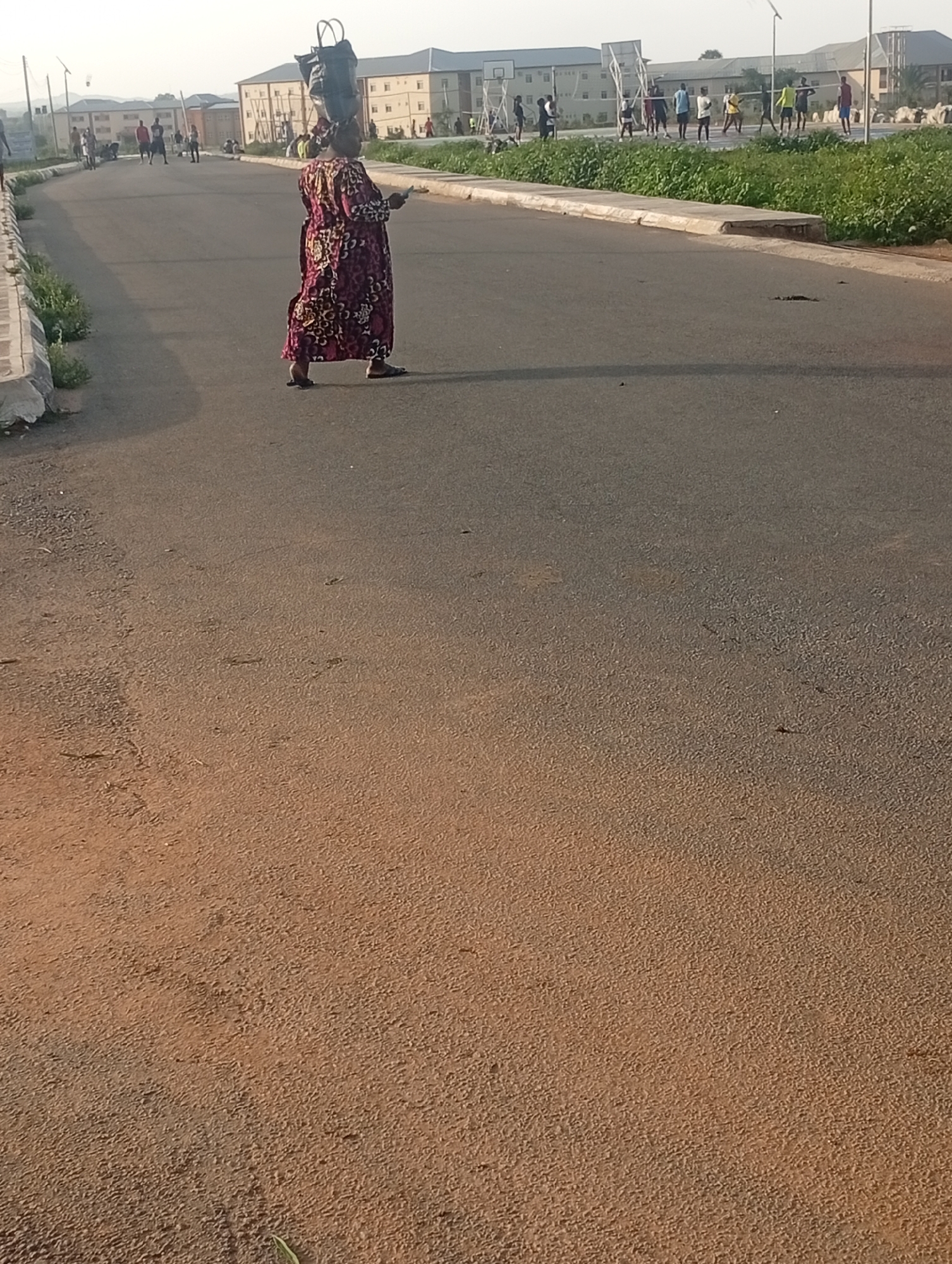 An outdoor, eye-level, full-shot image shows a woman balancing a large bundle of items on her head as she walks across an asphalt road. The woman is wearing a long, patterned dress in shades of red, yellow, and black. She is facing right, with her left hand holding a phone. Behind her, in the middle ground, several young people are playing a sport that appears to be volleyball on a court. In the background, there are several multi-story buildings, some with light-colored facades and dark roofs. The sky is bright and hazy. The scene takes place on a paved road with a dirt shoulder on the left, lined with green vegetation. The image appears to have been taken in the city of Yelwa, Nigeria.