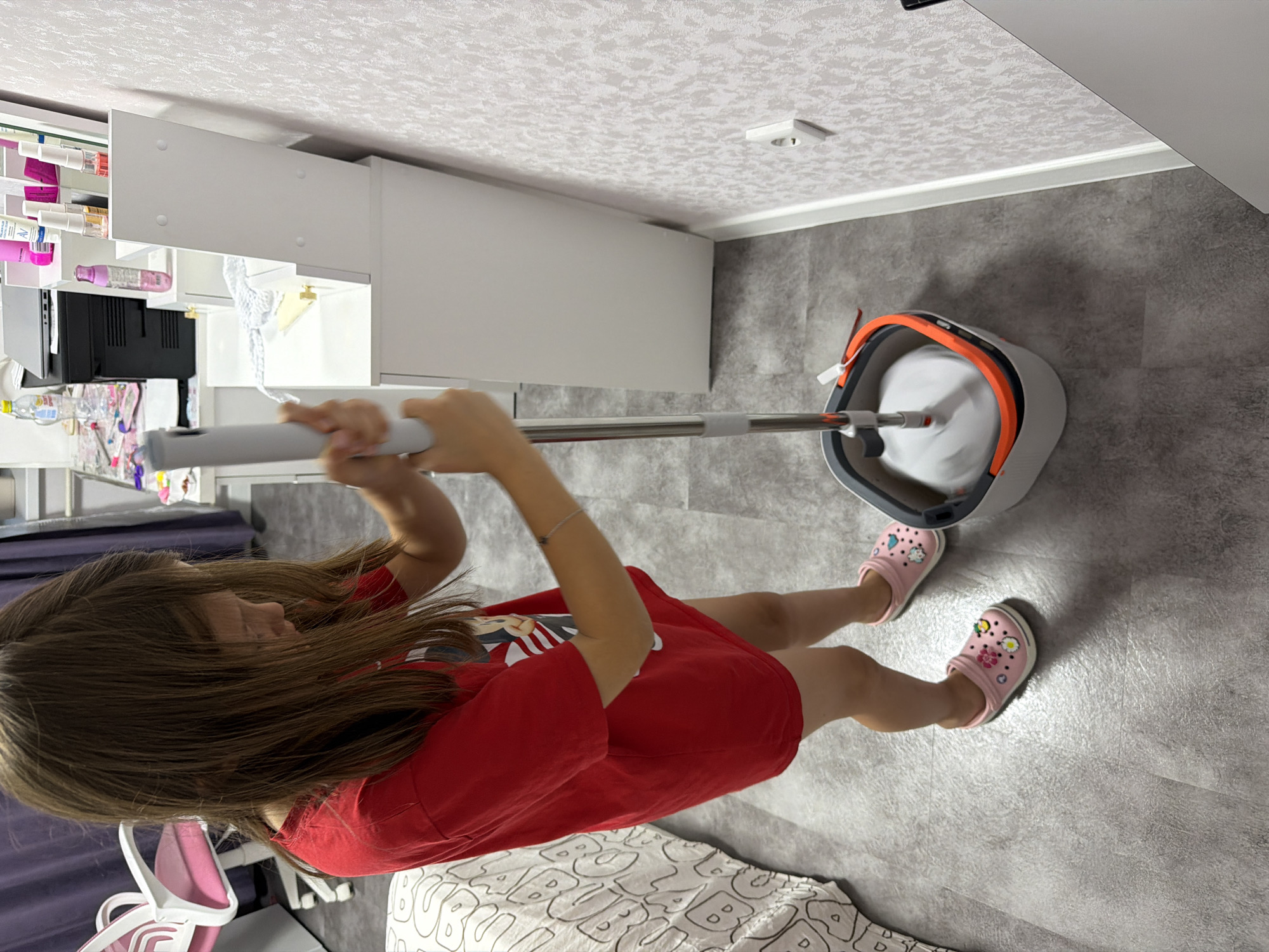 A young girl with long, brown hair, wearing a red t-shirt and pink crocs adorned with flowers and cartoon characters, is mopping a grey floor. She is holding a mop with a white circular head and an orange trim bucket. The mop is in motion, creating a blur. The background shows a white dresser or vanity with various bottles and cosmetics on top, a laptop, and a pink office chair with a white frame. The flooring appears to be laminate or linoleum, and the wall has a textured, light-colored wallpaper. The scene takes place indoors, likely in a bedroom or study area.