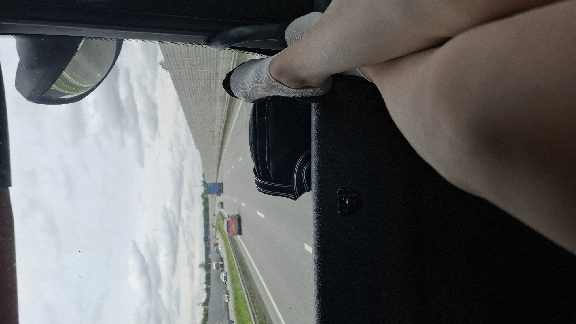 The image shows a person's legs and feet, wearing white socks with black trim, resting on a dark surface inside a vehicle. The person's legs are crossed, and their skin appears fair with some minor marks. They are positioned to look out the window, where a highway scene is visible.
The scene outside the vehicle is a multi-lane highway with other vehicles, including cars and a truck, traveling in the same direction. The highway is bordered by green grass and a concrete barrier. The sky is cloudy, suggesting overcast or partly cloudy weather. The perspective suggests the photo was taken from the passenger seat of a vehicle. There is a side-view mirror visible on the left side of the frame, reflecting the sky and a distant green area. On the dark surface in the foreground, there is a small black object with white text that reads "SOS" with a small asterisk.
The overall impression is a moment of travel, possibly on a road trip, with the focus on the relaxed posture of the passenger. The lighting indicates it is daytime. The specific mention of "Kurmanka, Russia" provides a geographical context for the location.