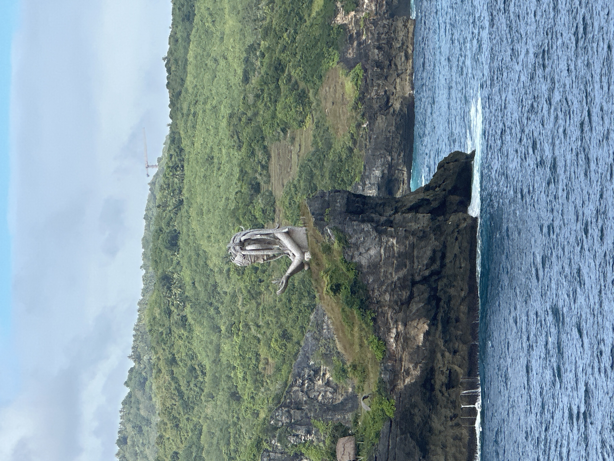 The image captures a stone statue of a woman with her right hand extended, located on a rock cliff on the island of Nusa Penida, Indonesia. Behind the statue is dense, green vegetation covering the hillside. The cliff faces the ocean, which occupies the right portion of the image. The sky is overcast. A construction crane is visible in the distance on the hillside.