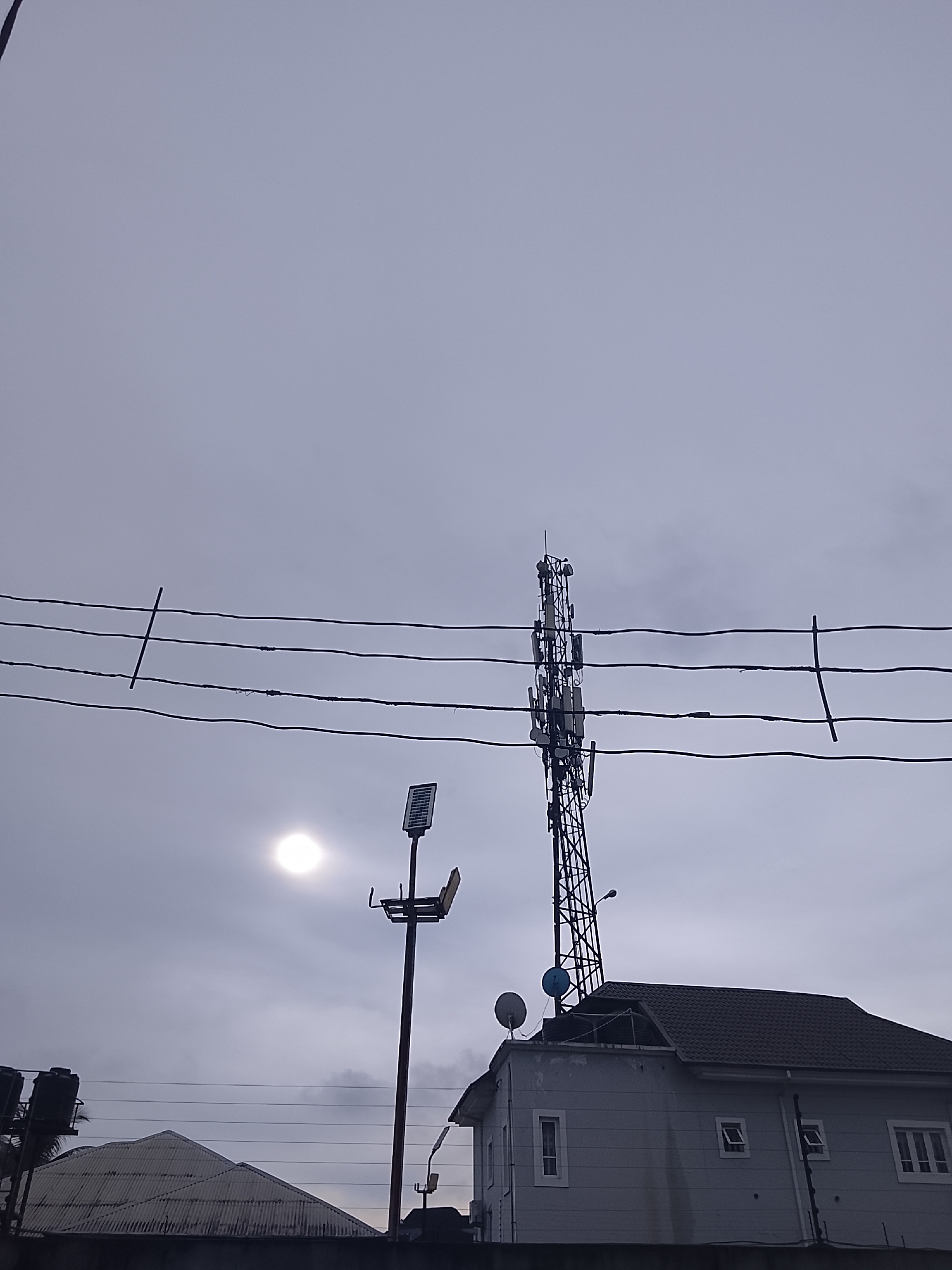 The image depicts an outdoor scene in Rumuepirikom, Nigeria, under an overcast sky. Dominating the frame is a tall cellular tower with multiple antennas and dishes mounted on it. In the foreground, a two-story building with a dark gray tiled roof is visible. The building has several windows, some of which are open. To the left of the building, a corrugated metal roof of another structure is visible. A street light with a solar panel is positioned to the left of the cellular tower, and a bright circular light source, likely the sun partially obscured by clouds, is present in the upper left portion of the image. Power lines are strung horizontally across the frame, supported by poles. The overall atmosphere is muted due to the cloudy conditions. There are no people visible in the image.