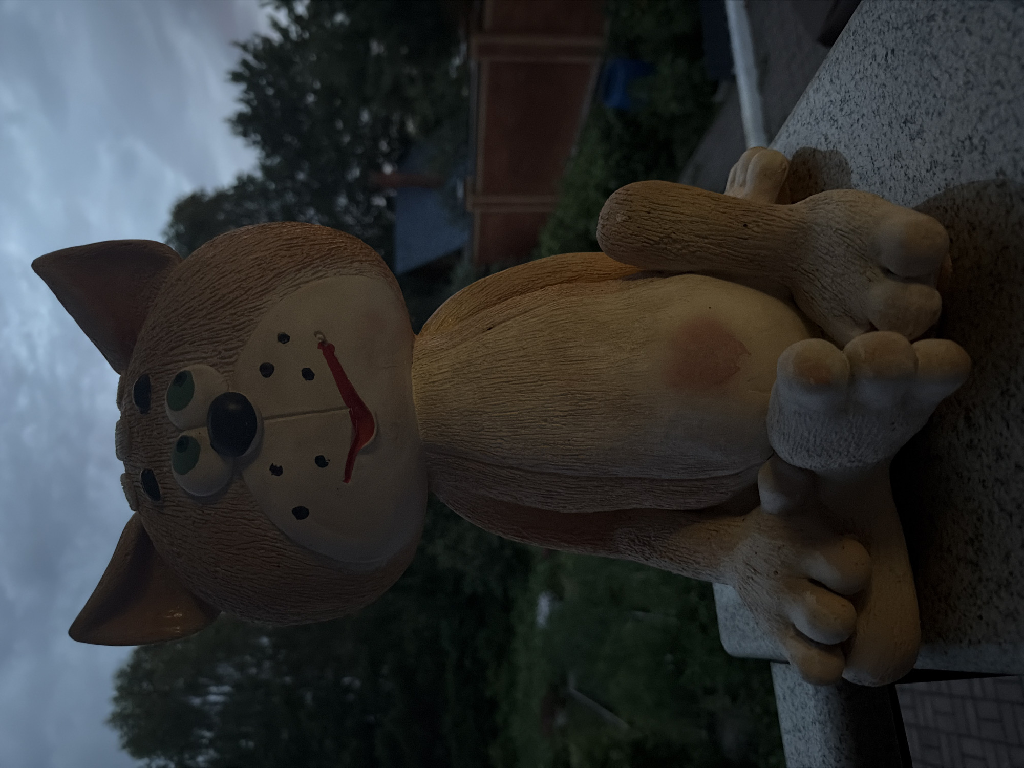 A ceramic cat figurine is seated on a stone ledge. The figurine is light brown with a cream-colored belly and paws. It has green eyes, a black nose, and a red mouth with black dots on its cheeks. The figurine is positioned outdoors, with greenery and trees visible in the background, along with a wooden fence and some structures. The sky is overcast and dark, suggesting it is either dusk or dawn. The overall scene appears to be a residential backyard or garden area, likely in the city of Kamenka, Russia, given the provided context.