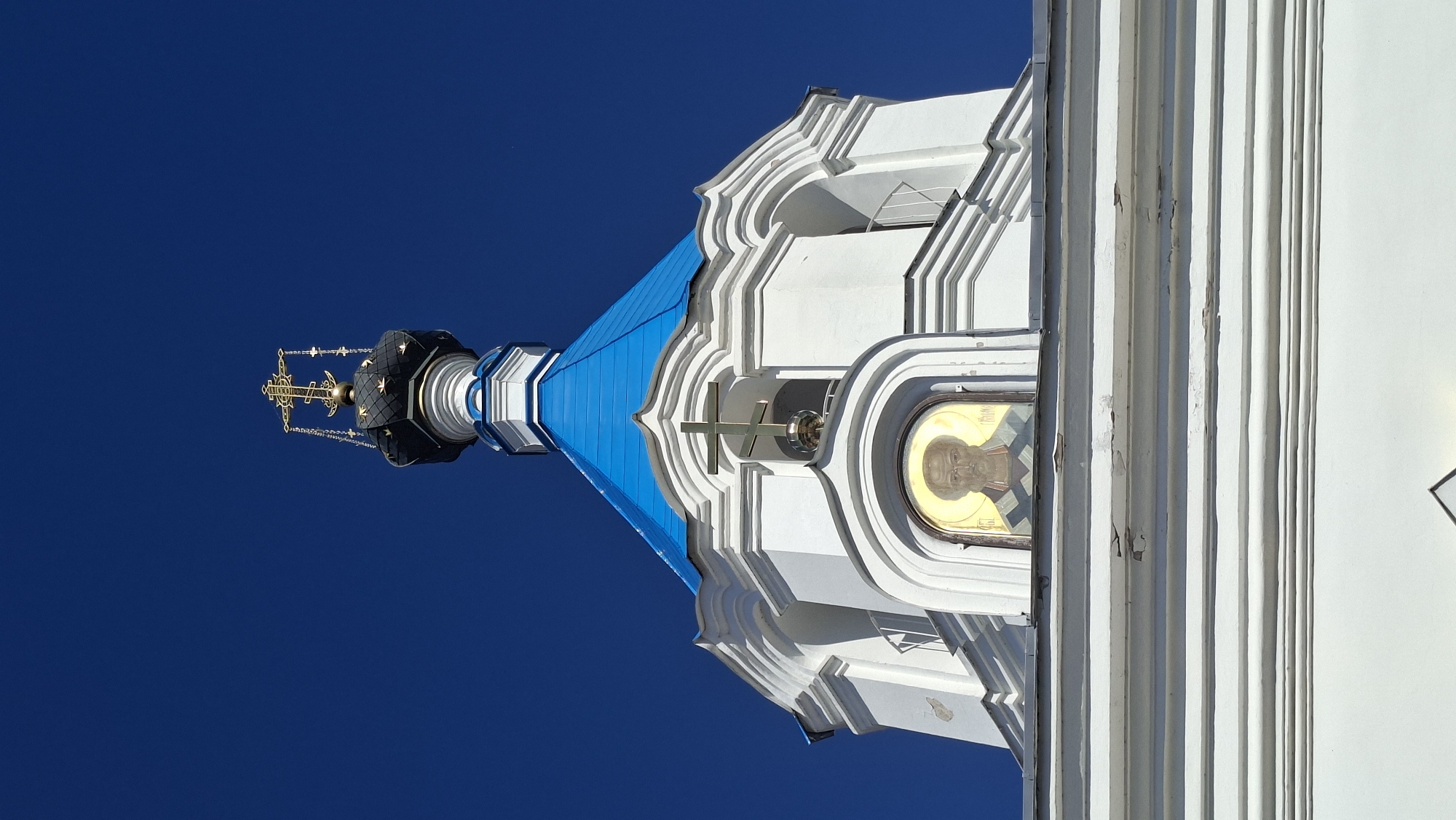 The image shows the top section of a white Orthodox church with a blue roof and a golden cross. An icon of a man with a beard, likely a saint, is visible in an alcove beneath the cross. The church is against a clear blue sky. The scene is a detail shot of architectural and religious elements, not a wide view of the city of Pastavy, Belarus.