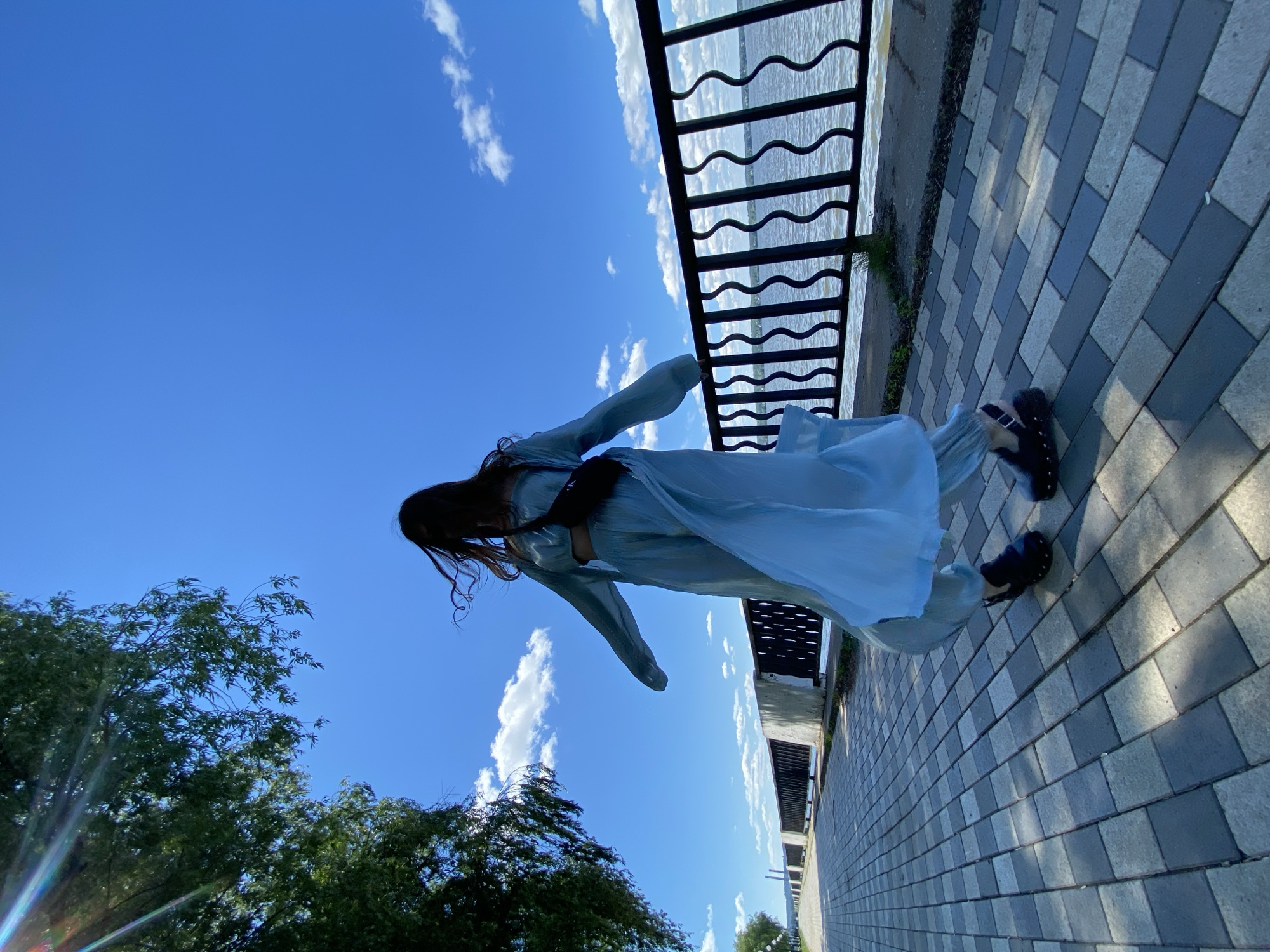 A woman is pictured outdoors in a bright, sunny environment. She is wearing a flowing light blue outfit that appears to be a set with a top and wide-leg pants, and black platform sandals. A dark bag is slung across her body. Her hair is long and dark, and it is being blown by the wind, partially obscuring her face. She is standing on a paved walkway made of rectangular gray and black stones. To her right, there is a black metal fence with a wavy pattern, beyond which lies a body of water. In the background, there is a line of trees on the left side of the walkway, and the sky is a clear, vibrant blue with scattered white clouds. The sun is casting a lens flare into the image from the upper left.
