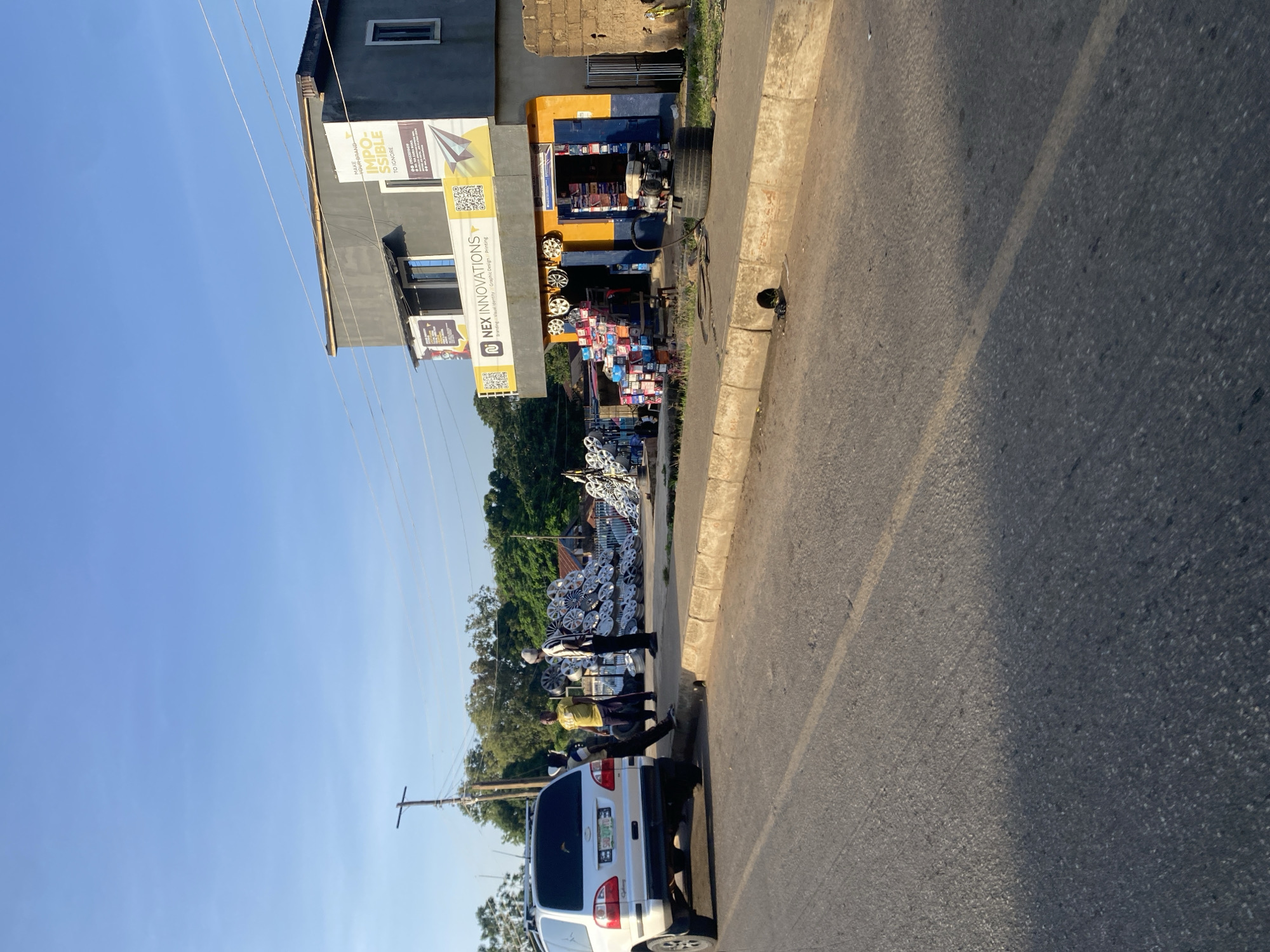 In New Stadium, Nigeria, a bright, clear sky stretches above a street-side business. The establishment, "NEX INNOVATIONS," is housed in a two-story building with a grey upper level and a vibrant orange and blue storefront. Displayed prominently on the building are advertisements for branding, visual identity, graphic design, and printing services.
In front of the store, a collection of car wheels are neatly stacked, suggesting the business may also be involved in automotive services or sales. A white SUV is parked on the side of the road, partially obscuring the view of some of the stacked wheels. Several individuals are present in the scene. Two men stand near the stacked wheels, engaged in conversation or work. Another individual is positioned near the white SUV.
The environment suggests a commercial area with active street life. The road surface is paved, and a concrete curb separates the road from the sidewalk where the business and displayed items are located. The background features a line of trees and other buildings, indicative of a populated urban setting. The lighting suggests it is daytime, possibly late afternoon given the angle of the shadows.