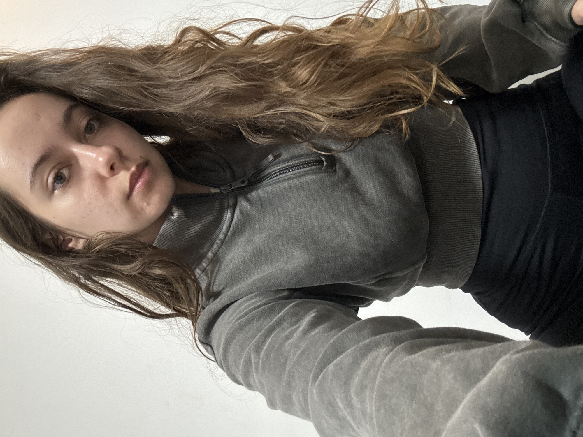 A young woman with long, wavy brown hair is facing the camera. She is wearing a gray, zip-up sweatshirt and black leggings. The background is a plain white wall. It is not possible to ascertain the location of the image as Ramat Gan, Israel based on the provided information.