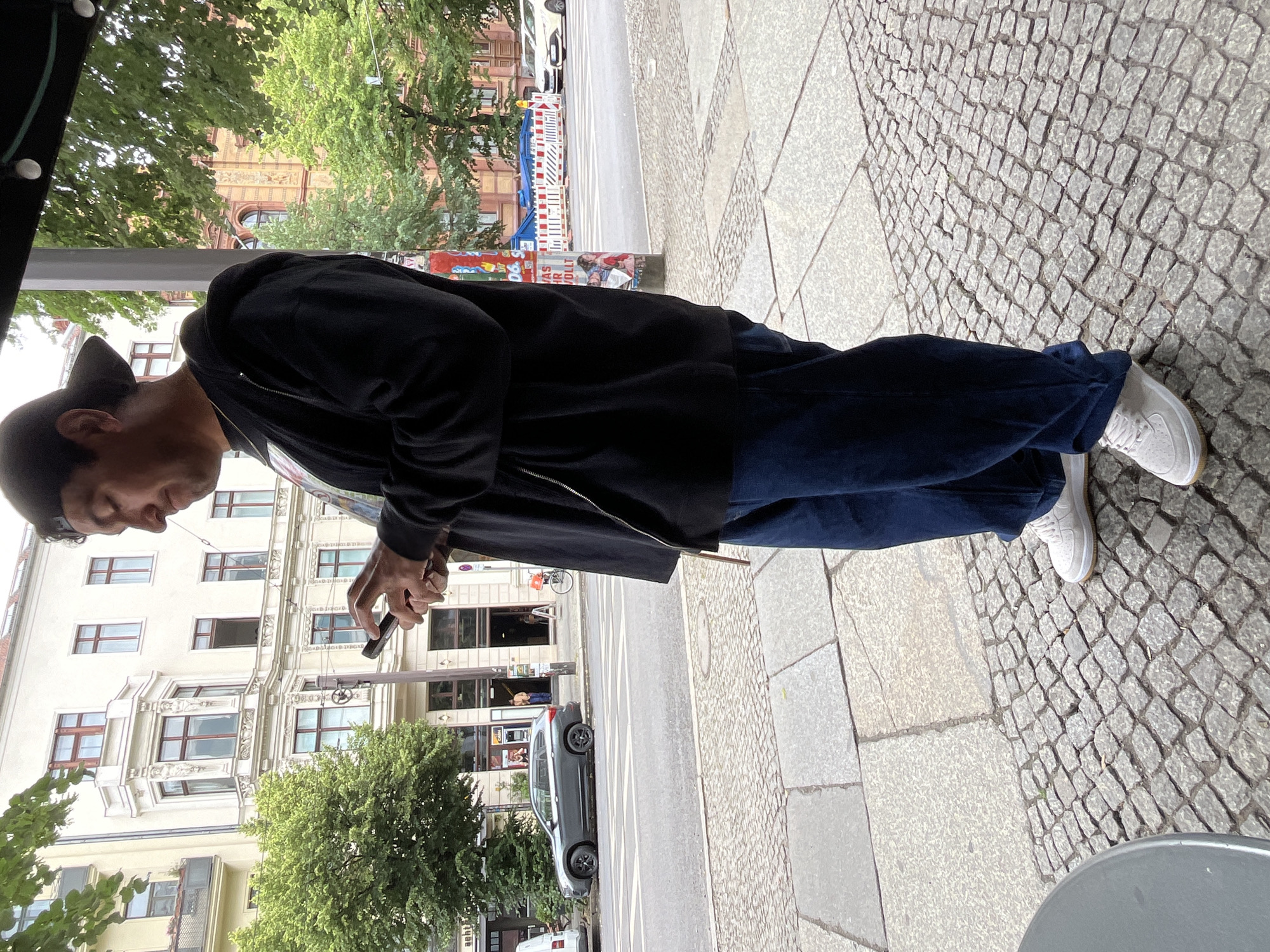 A man wearing a black baseball cap, black zip-up hoodie, and dark blue jeans is standing on a cobblestone sidewalk, looking down at his phone. He is holding the phone with both hands. The man is in the foreground, and his body is angled to the right. Behind him, across a street, is a building with multiple windows and a decorative facade. There are trees lining the street and a dark grey car parked on the side. Further down the street, there are other buildings and a construction site with barriers. The sky is overcast. The overall setting suggests an urban environment in Berlin, Germany.