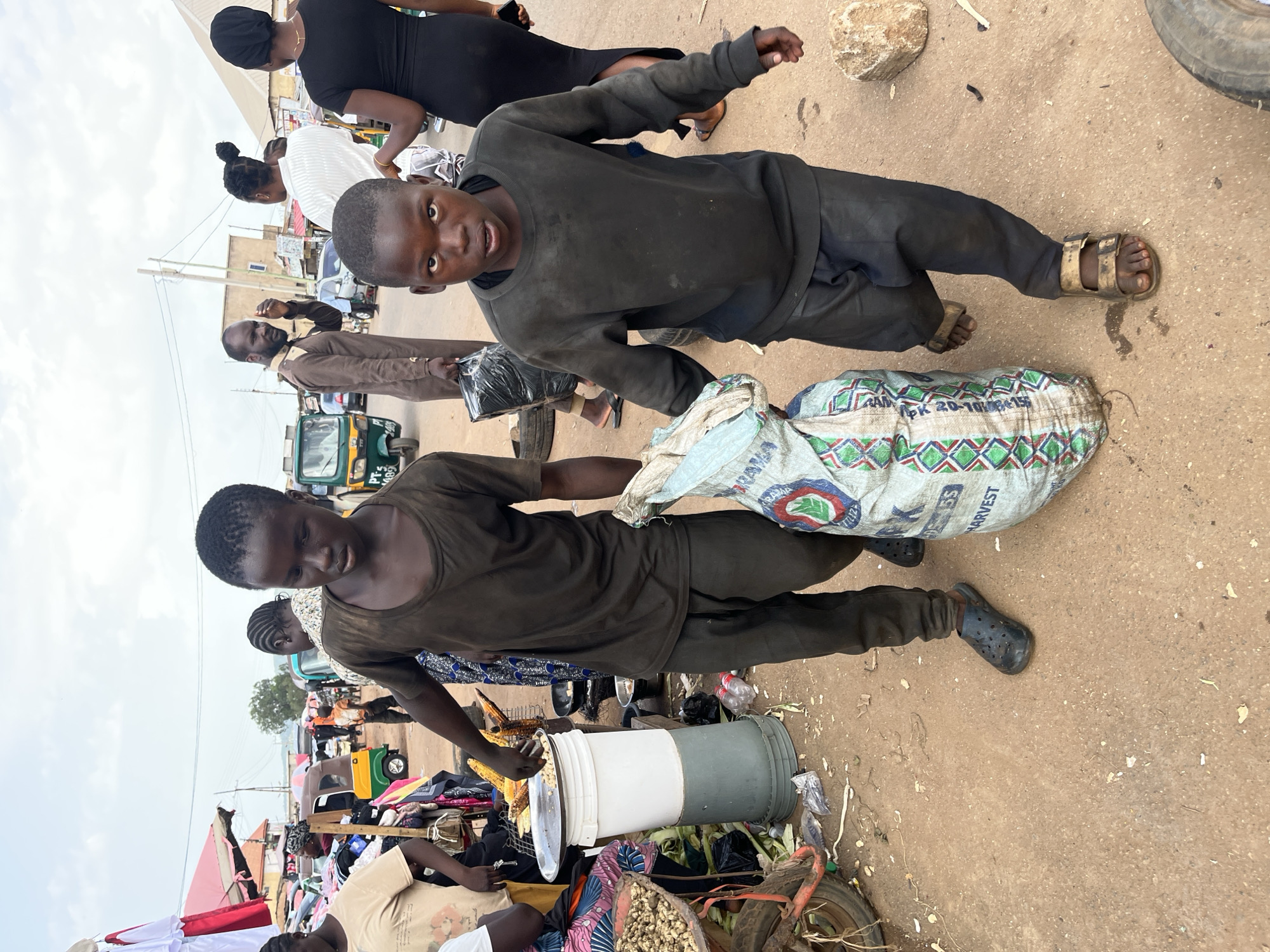 In Rafin Par, Nigeria, an outdoor market scene unfolds under what appears to be a cloudy sky. The foreground is dominated by two young boys, their faces turned towards the camera. The boy on the left wears a dark t-shirt and dark pants, looking down with a neutral expression. Beside him, another boy in a dark long-sleeved shirt and dark pants gazes directly at the viewer, his expression one of curiosity. He is positioned near a large, patterned sack with the words "HARVEST" visible, suggesting agricultural produce.
Behind them, other individuals are present. A woman in a black headscarf and black dress stands with her back to the camera, while another woman in a white striped shirt is partially visible. Further back, a man in a brown jacket is seen conversing. A prominent object in the scene is a green and yellow auto-rickshaw, commonly known as a "keke," which suggests a mode of local transportation.
The ground is a dusty, earthy surface, typical of an outdoor market. Scattered items like plastic bags, what appear to be vegetables, and a large white and grey cylindrical container are visible, indicating the presence of vendors and shoppers. The overall atmosphere suggests a busy, everyday market environment. The composition of the image, with the subjects prominently featured and looking towards the camera, creates a direct connection with the viewer.