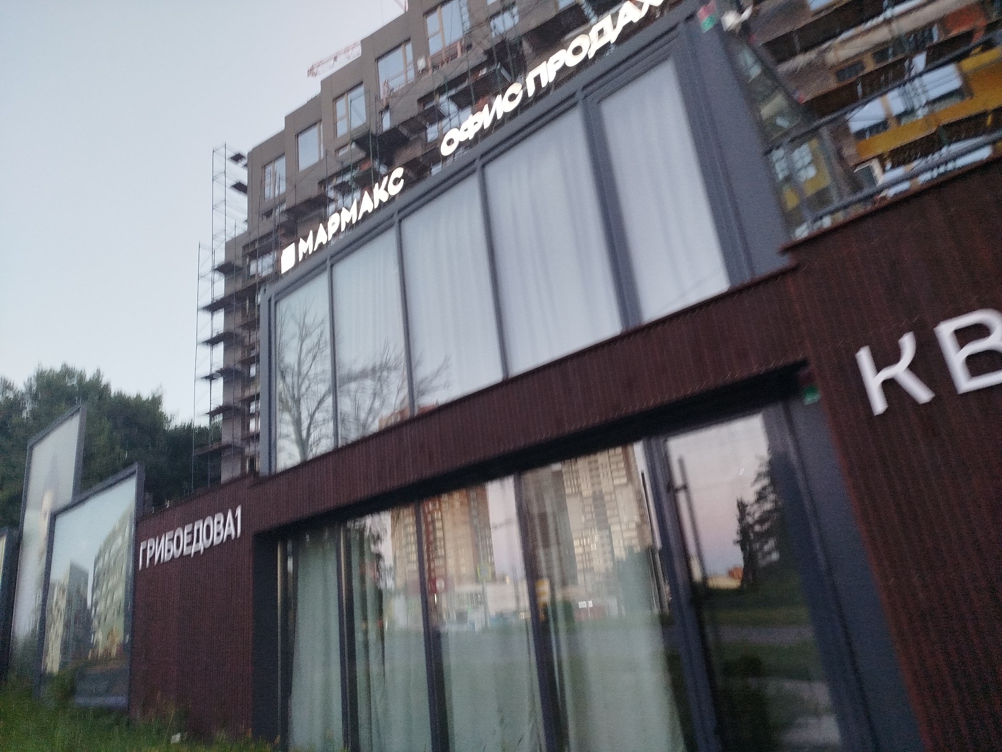 The image shows a modern building with large windows and a dark brown, textured facade. A white sign with Cyrillic text is mounted on the building. The text reads "ГРИБОЕДОВА1" on the left side of the building. On the upper part of the building, another white sign reads "МАРМАКС ОФИС ПРОДАЖ". The building appears to be under construction, as indicated by scaffolding on the side and the unfinished state of the upper floors of the adjacent structure. Reflections of buildings and trees are visible in the windows. The overall scene suggests a commercial or residential development project in an urban setting. The location is identified as Ryazan, Russia.