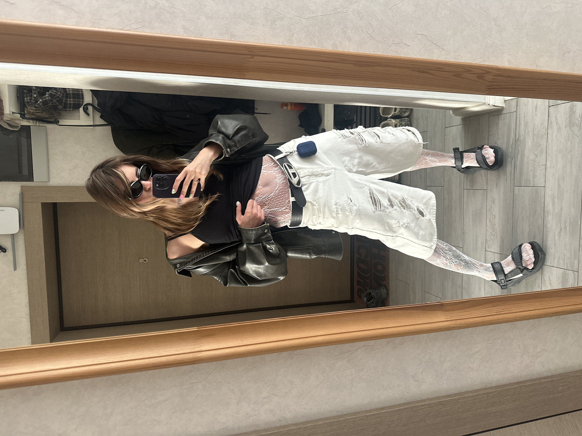 A young woman stands in front of a full-length mirror, taking a selfie. She is dressed in a black tank top, a dark leather jacket, distressed white denim shorts, and black sandals. She wears white fishnet stockings. Her hair is brown and shoulder-length, and she has on sunglasses. A phone is held up to take the picture. The mirror is framed by light wood, and the room appears to be an entryway or hallway, with a closed door in the background and a coat rack with jackets visible to the right. The floor is tiled. The image was taken indoors, likely in an apartment.