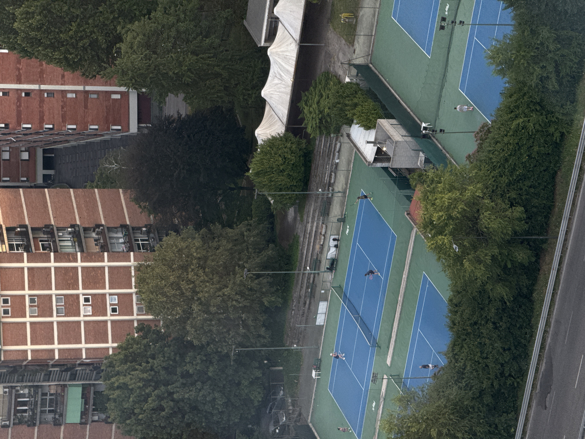 The image shows an aerial view of several outdoor tennis courts with blue surfaces and green surrounds, likely located in Milan, Italy. Some of the courts have nets and are illuminated by tall light poles. There are several people on the courts, some appearing to be playing tennis, while others are standing or walking. In the background, there are tall brick buildings with many windows, characteristic of apartment complexes. Lush green trees surround the tennis courts, and a road with a metal barrier runs along the bottom of the frame. A few parked cars are visible near a fenced area on the left side of the image. There are also white tent-like structures in the middle ground, possibly used for shade or events. The overall scene suggests an urban recreational area integrated with residential buildings and natural greenery.