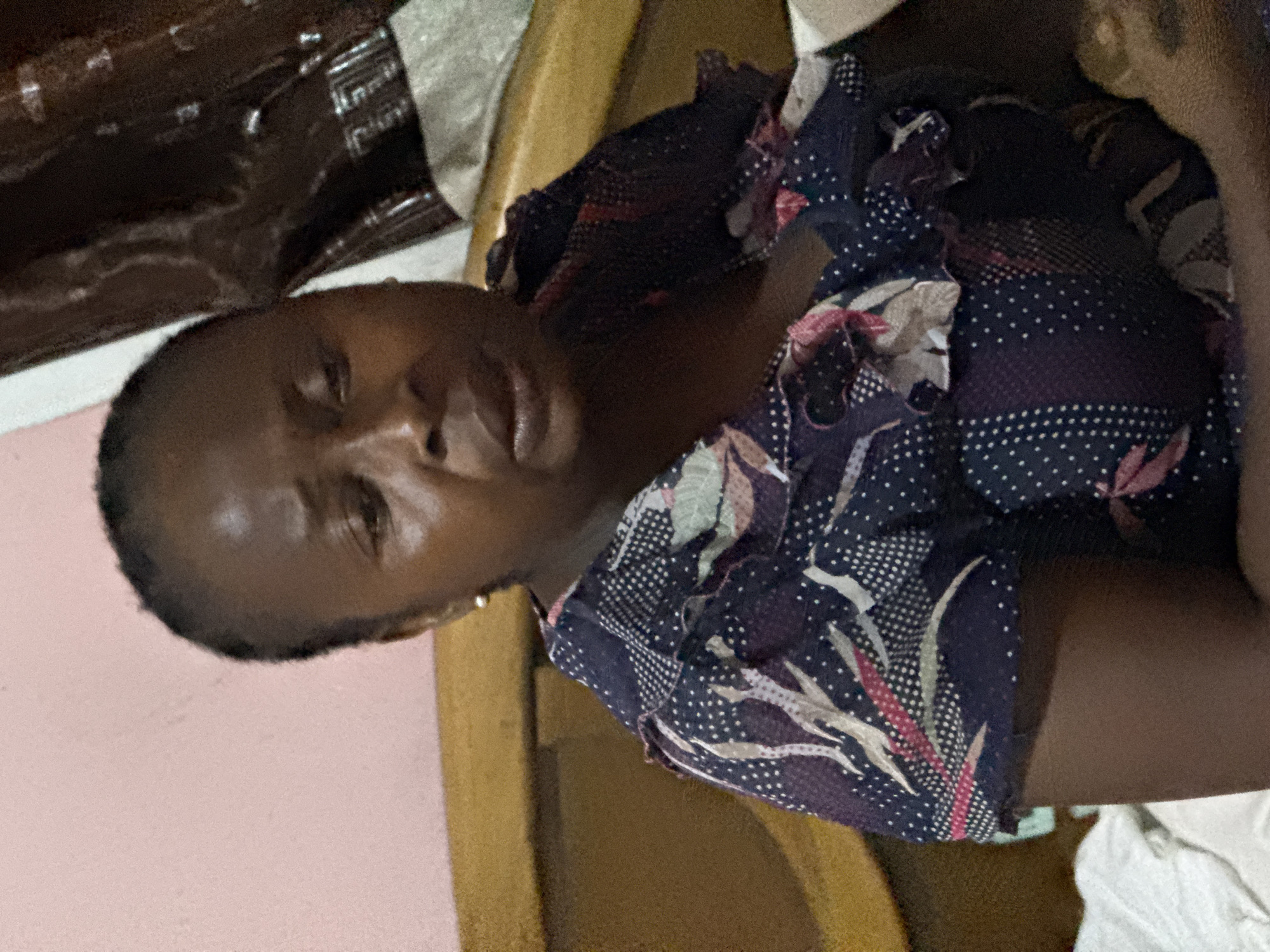 A woman is seated in an indoor environment. She is wearing a dark blue and pink blouse. The location appears to be in a residential setting, possibly in Abuja, Nigeria, although there is no definitive visual confirmation of the city. The environment includes a wooden structure, possibly a chair or part of the room's decor. Other items are also visible, including fabric.