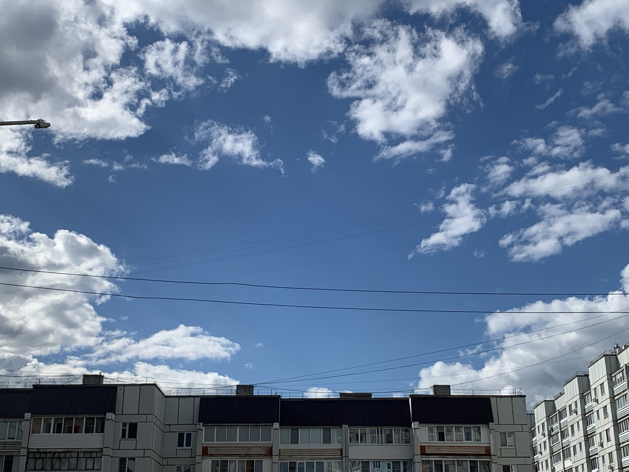 The image depicts a sky with scattered white cumulus clouds against a clear blue sky. In the foreground, the rooftops and upper floors of several multi-story apartment buildings are visible. These buildings appear to be located in an urban environment, likely Chaikovsky, Russia. Overhead, a network of electrical wires crisscrosses the frame. A street light is visible on the left side of the image. The architecture of the buildings suggests a Soviet-era or post-Soviet construction style, common in many Russian cities. The scene is captured from a low angle, looking upwards towards the sky and the tops of the buildings.
