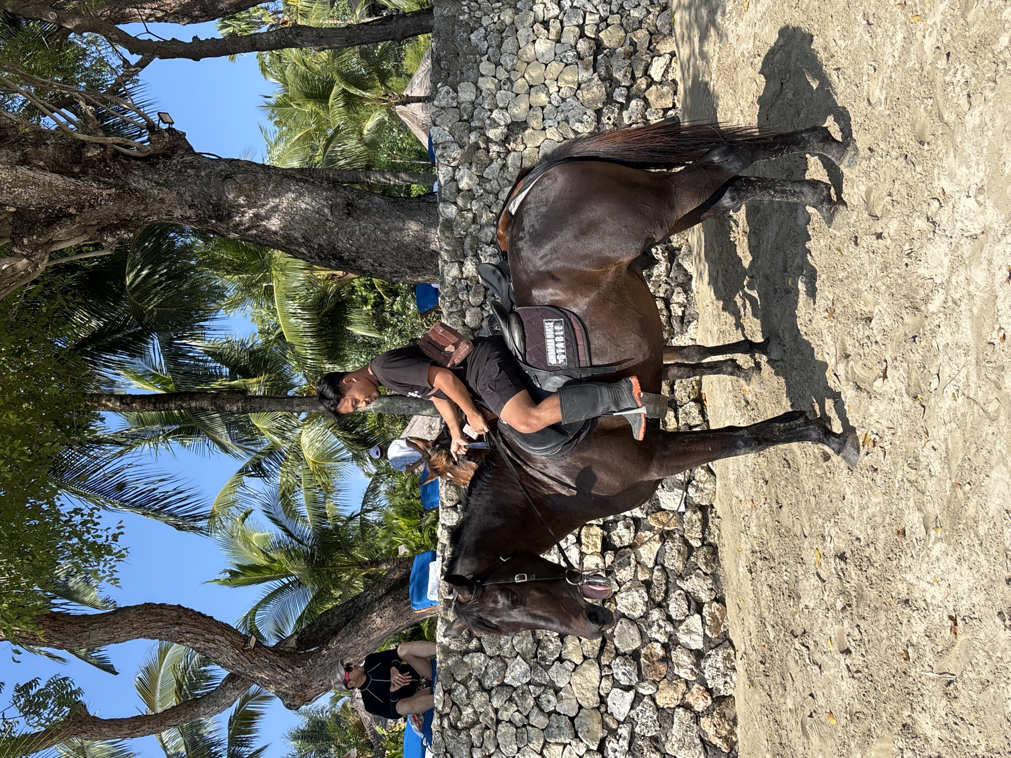 An Indonesian man in a black t-shirt and shorts rides a dark brown horse on a sandy path in Kuta Selatan, Indonesia. The man wears tall black boots with an orange stripe. The horse has a saddle with a pad that reads "NUSA DUASA HORSE STABLE". Another horse is partially visible behind the first, with its rider wearing a white shirt and a cap. In the background, a rough stone wall runs horizontally, topped with green foliage and palm trees. Further back, two people are seated on blue mats under the shade of a large tree. One person wears a black long-sleeved shirt, sunglasses, and a cap, while the other is partially obscured. The scene is under a clear blue sky, suggesting it is daytime and sunny, with the sandy ground showing some fallen leaves.