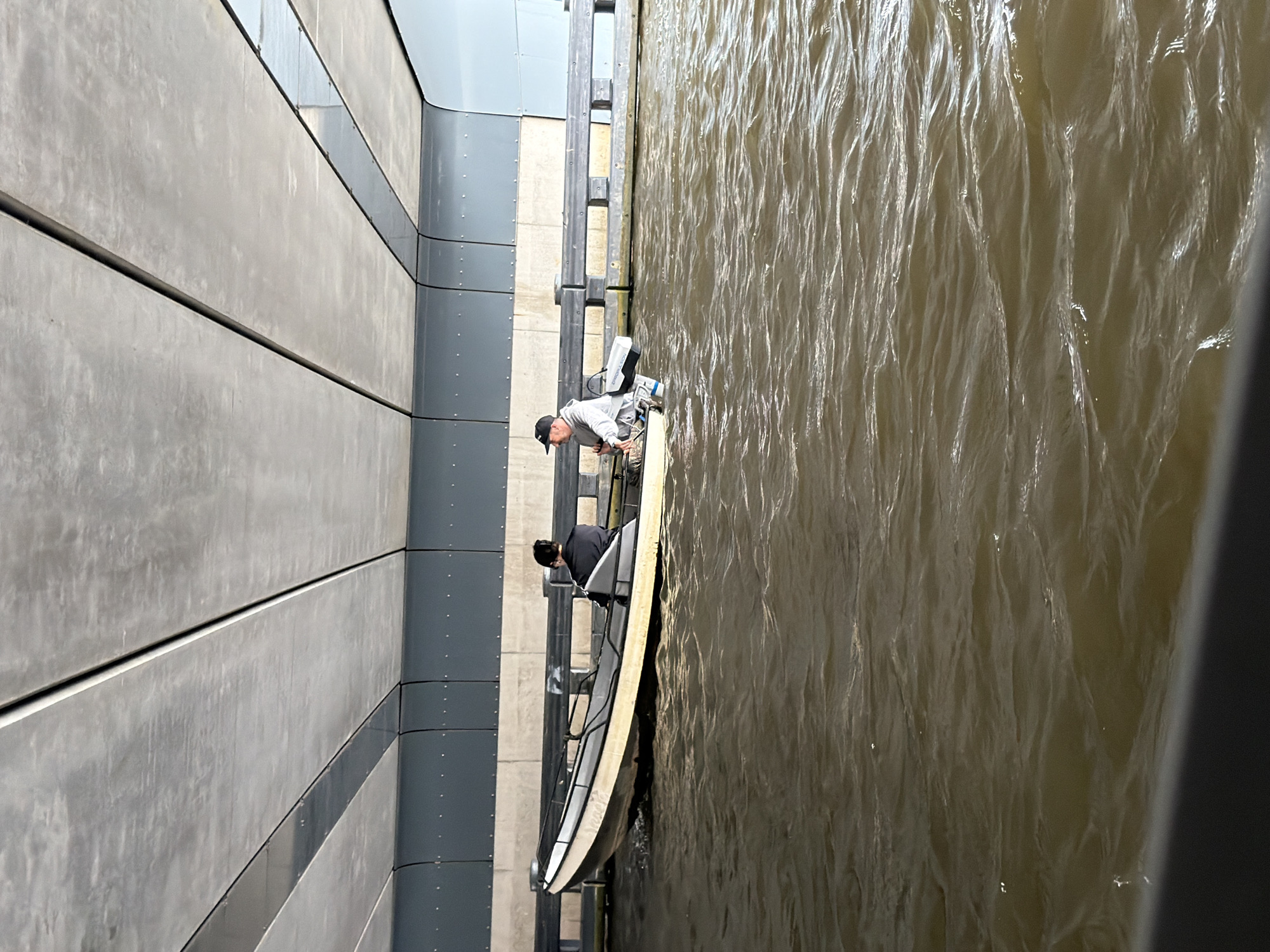 Two individuals are in a small boat, navigating through flooded waters under a concrete overpass in Amsterdam, Netherlands. The water is murky and choppy, with visible waves. The boat appears to be a jon boat or a similar small utility craft, equipped with a small outboard motor. One person, wearing a dark shirt, is seated towards the front of the boat, facing away from the camera. The other person, wearing a light-colored hoodie and a baseball cap, is seated towards the rear of the boat, facing the camera, and appears to be operating the motor. A bag is visible in the boat near the rear passenger. The overpass above is constructed of large concrete slabs, with a gray metal structure running along the sides. The context suggests a flood event, with normal infrastructure submerged.