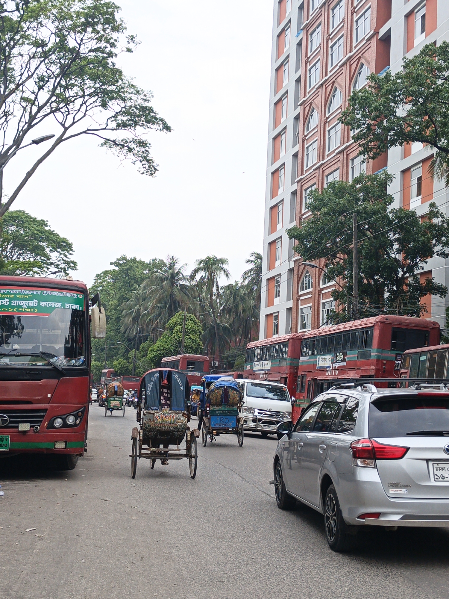 The image shows a street scene in a city in Bangladesh. The street is filled with vehicles, including buses, rickshaws, and a silver car. The buses are red and have writing in Bengali on them. Rickshaws are in the center of the road. The silver car is parked on the side of the road and has a license plate. There is a tall building in the background with a red brick facade. Trees line the street. The sky is overcast. The writing in Bengali on the bus to the left includes text indicating the bus is associated with "East Graduate College, Dhaka." The registration plate of the silver car says "Dhaka."