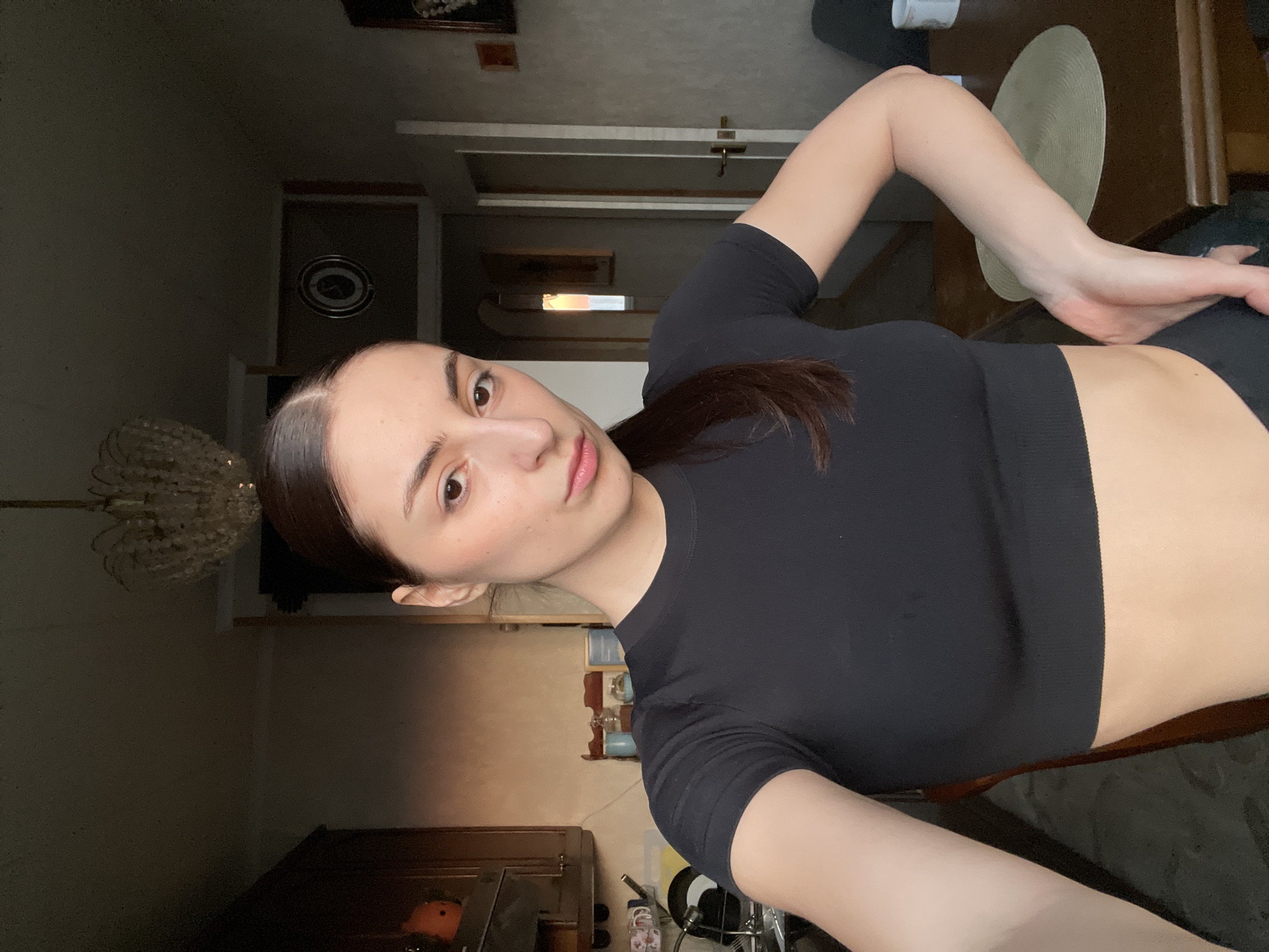 A young woman is depicted in a residential apartment in Moscow, Russia. The subject, with dark brown hair pulled back into a sleek style, wears a black, short-sleeved crop top and dark bottoms. She is positioned facing the camera, extending her right arm, suggesting a selfie, with her left hand placed on her hip. Her expression is neutral, and her gaze is directed forward.
The background reveals an interior space that functions as both a kitchen and dining area. To the subject's left, dark wooden kitchen cabinets are visible above a white sink with a faucet. Several dishes, including mugs and plates, are present around the sink. A small, orange decorative item rests on a counter or shelf. The wall in this section is light beige.
To the subject's right, a dark wooden dining table holds a circular, light-colored woven placemat and a white mug. A dark upholstered chair or sofa is partially visible behind the table. A light-colored door, slightly ajar, leads into an adjacent room or hallway, revealing an arched doorway and a brightly lit window in the distance, indicating natural light ingress. A dark-framed circular object, possibly a clock or mirror, is mounted on the wall above the door, and two framed pictures hang further to the right. A multi-tiered crystal chandelier descends from the ceiling in the upper center of the frame. The overall lighting in the immediate area is ambient, supplemented by natural light from the distant window.