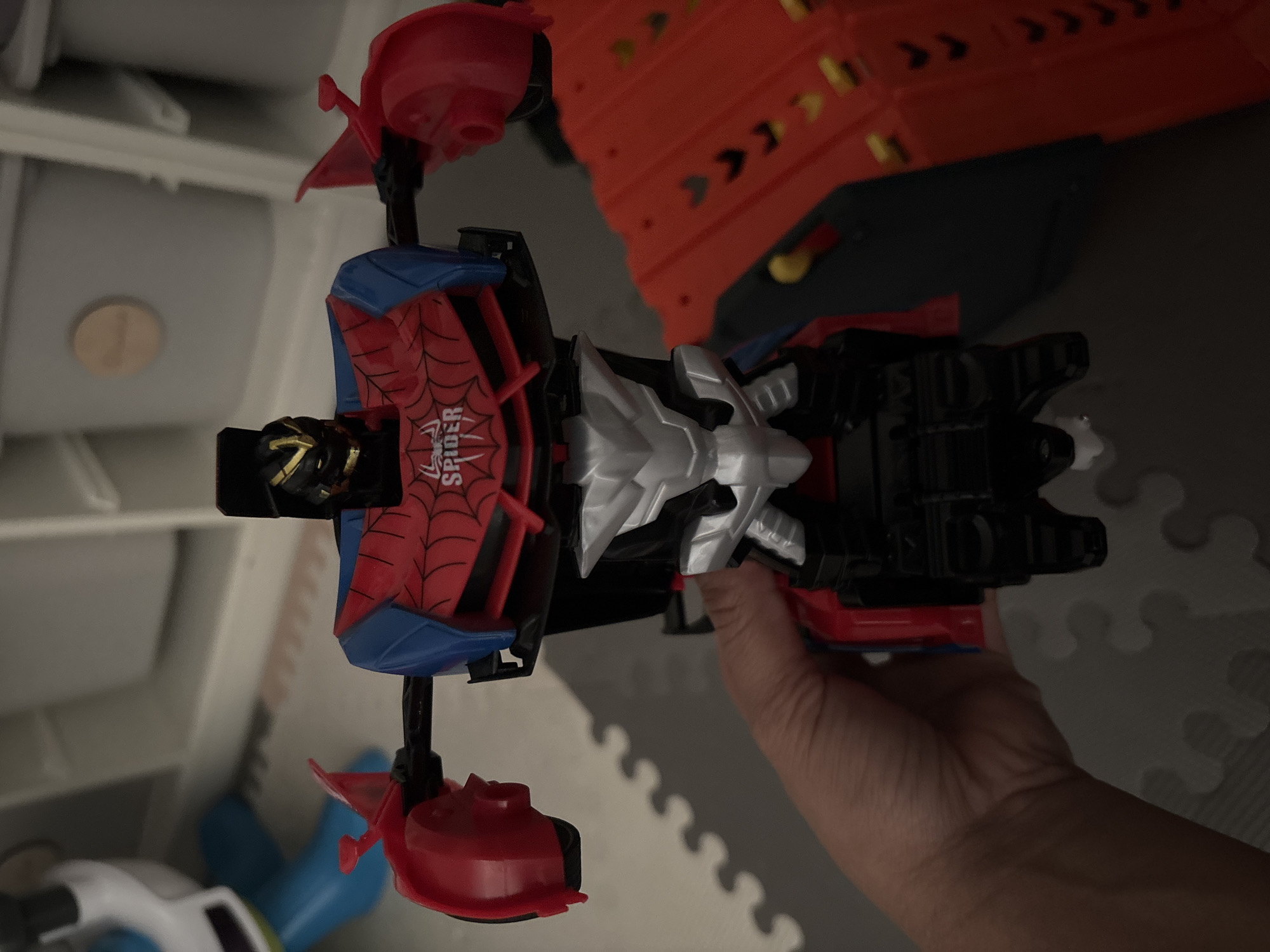 A human hand holds a red, blue, black, and silver transforming robot toy featuring a black head with a gold visor and a chest emblazoned with a spiderweb design and the text 'SPIDER'. The toy's arms are extended outwards, with red sections resembling wheel wells. The scene is indoors, on a grey interlocking foam play mat, visible at the bottom and left. In the blurred background to the left, a blue and white plastic toy is partially visible. To the right, a red toy car track with black chevron markings and yellow details is partially seen. The upper background reveals white shelving units holding light grey storage bins, one with a circular wooden tag attached. No specific visual cues within the image indicate a location in the city of Dubai, United Arab Emirates.