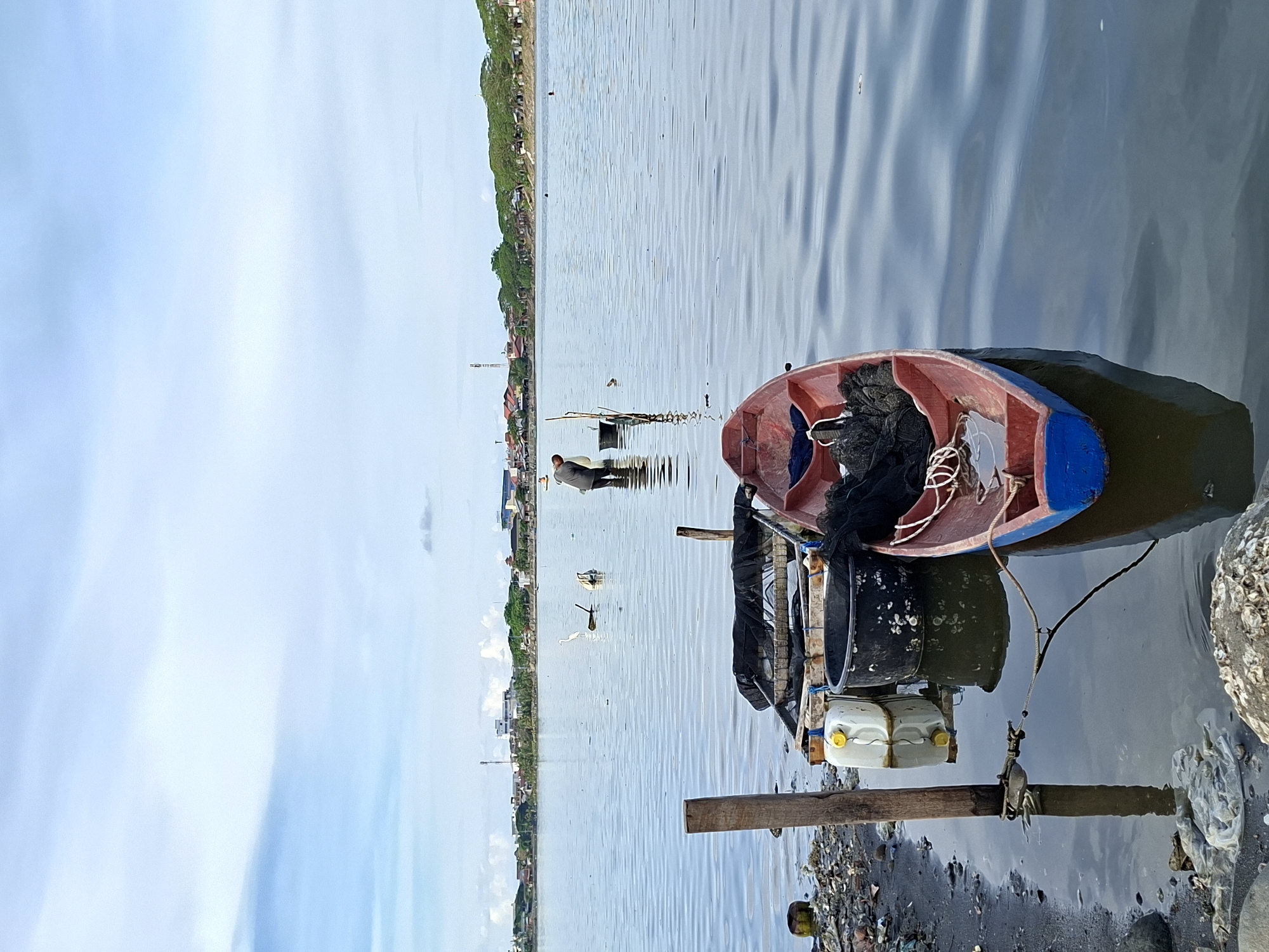 The image shows a small boat docked in a body of water, likely a lake or harbor. The boat is red and blue, with fishing nets inside. A wooden post is used to secure the boat near the shoreline. In the distance, there appears to be a person standing in the water. Several white birds are also visible in the water. The shoreline is polluted with plastic and trash. In the background, a populated area with buildings and trees suggests the location is near a city. Considering the image's context, this could be a scene from Lhokseumawe, Indonesia, possibly near the coast where fishing activities are common.