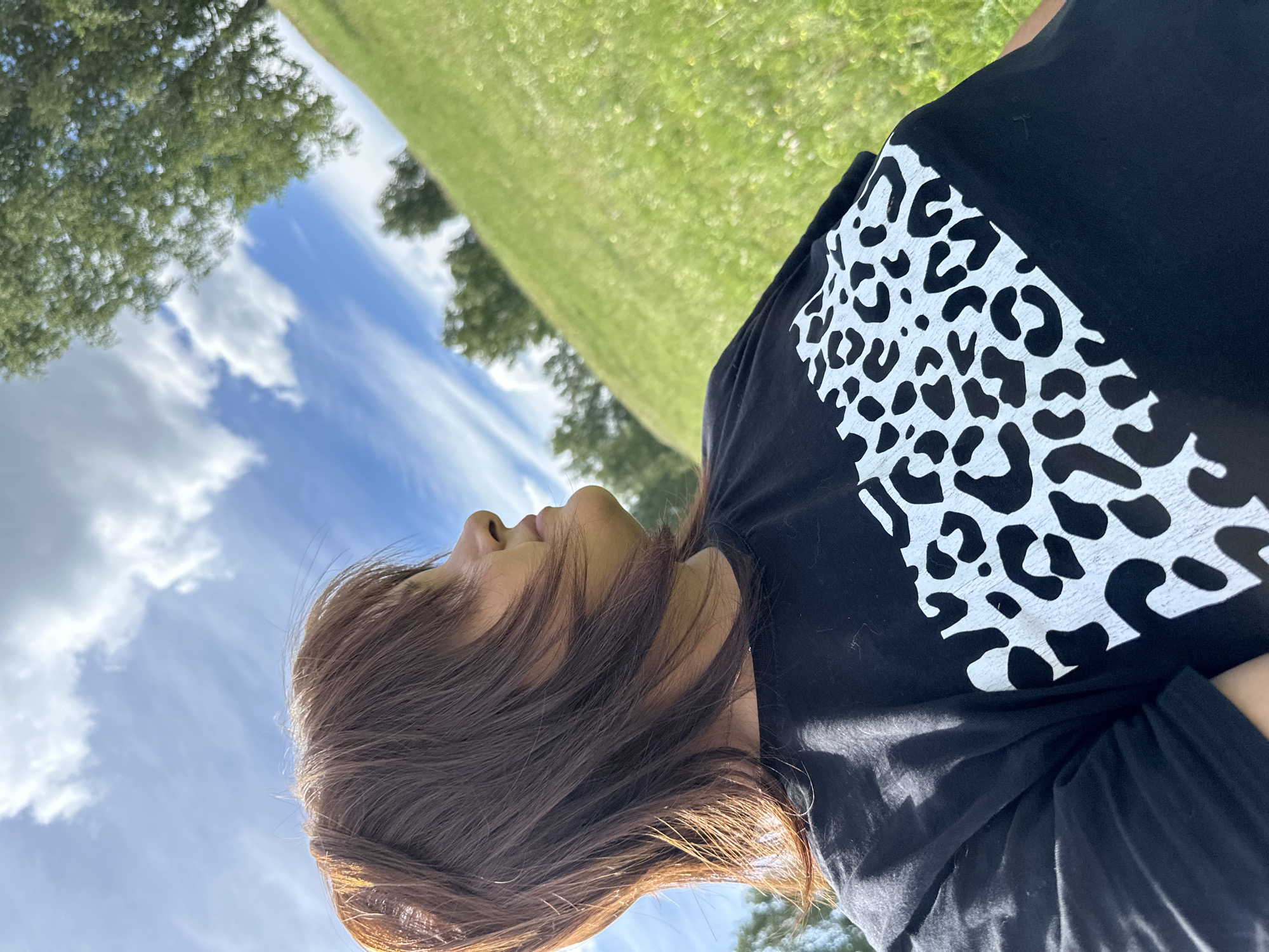 An outdoor, low-angle, medium shot captures a woman from the chest up, looking to her right with her head tilted upwards, as she stands in a grassy field under a bright, partly cloudy blue sky. She has medium-length, dark brown hair with bangs, and she is wearing a black t-shirt with a white leopard print design across the chest. The background is a green, sloping field with trees visible in the distance, and the sky is filled with scattered white clouds and wispy cirrus formations. The sun casts light from the right side of the frame, highlighting the woman's hair and the texture of the grass.