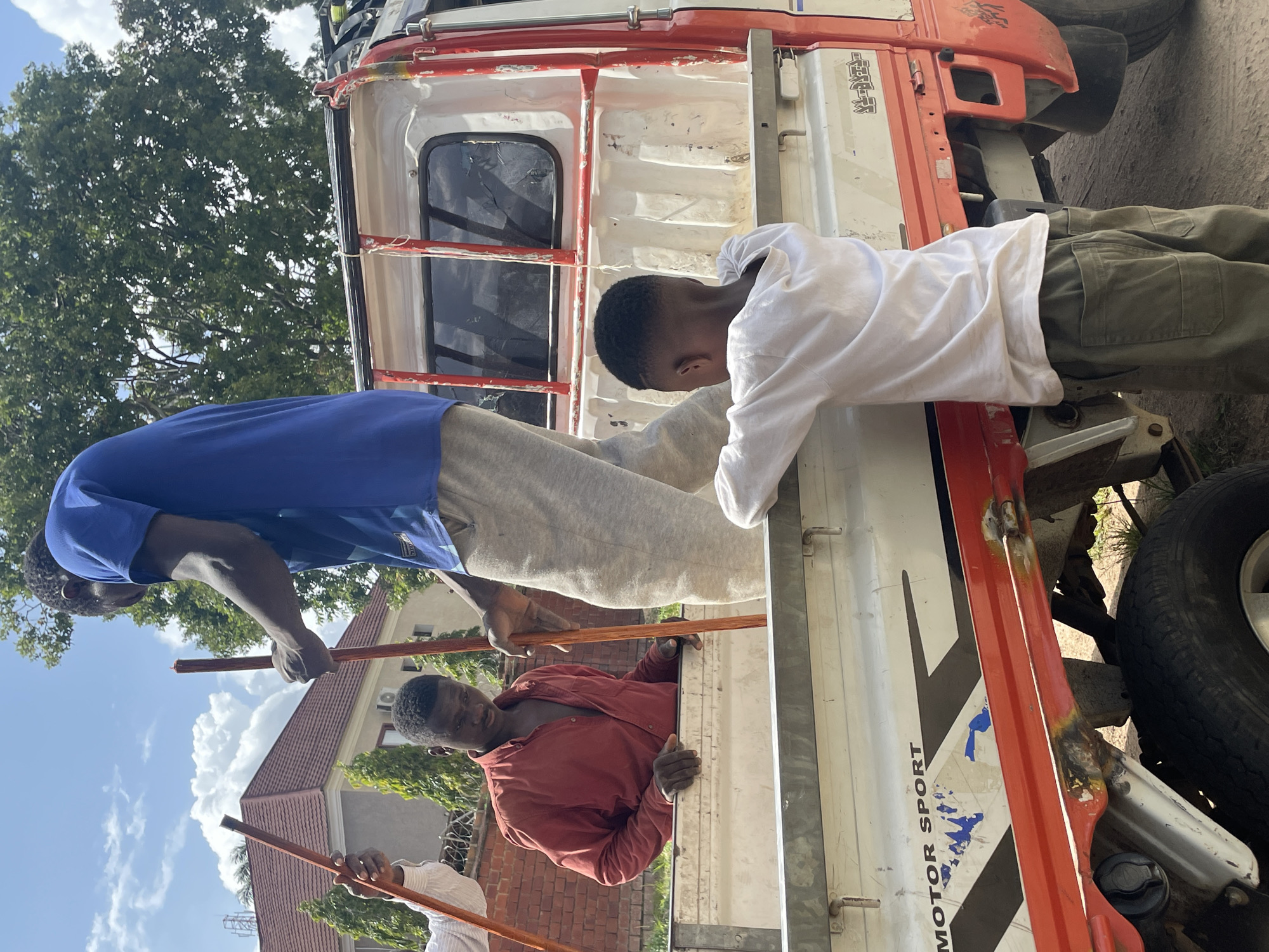 The image captures an outdoor scene in the city of Jos, Nigeria, depicting several individuals interacting with a pickup truck. In the foreground, a young boy wearing a white long-sleeved shirt and green cargo pants leans over the edge of the truck bed, his back to the camera. To his left, a man in a red collared shirt is also positioned within the truck bed, looking downwards. Another man, wearing a blue t-shirt and grey sweatpants, stands taller in the truck bed, holding a wooden pole. A third person, partially visible on the far left, also holds a similar wooden pole. The truck itself is white with red accents and appears to be a flatbed model. The background reveals a building with a red tiled roof, trees, and a partly cloudy sky. The ground beneath the truck appears to be dirt or gravel. The overall impression is one of a collaborative activity involving these individuals and the truck.