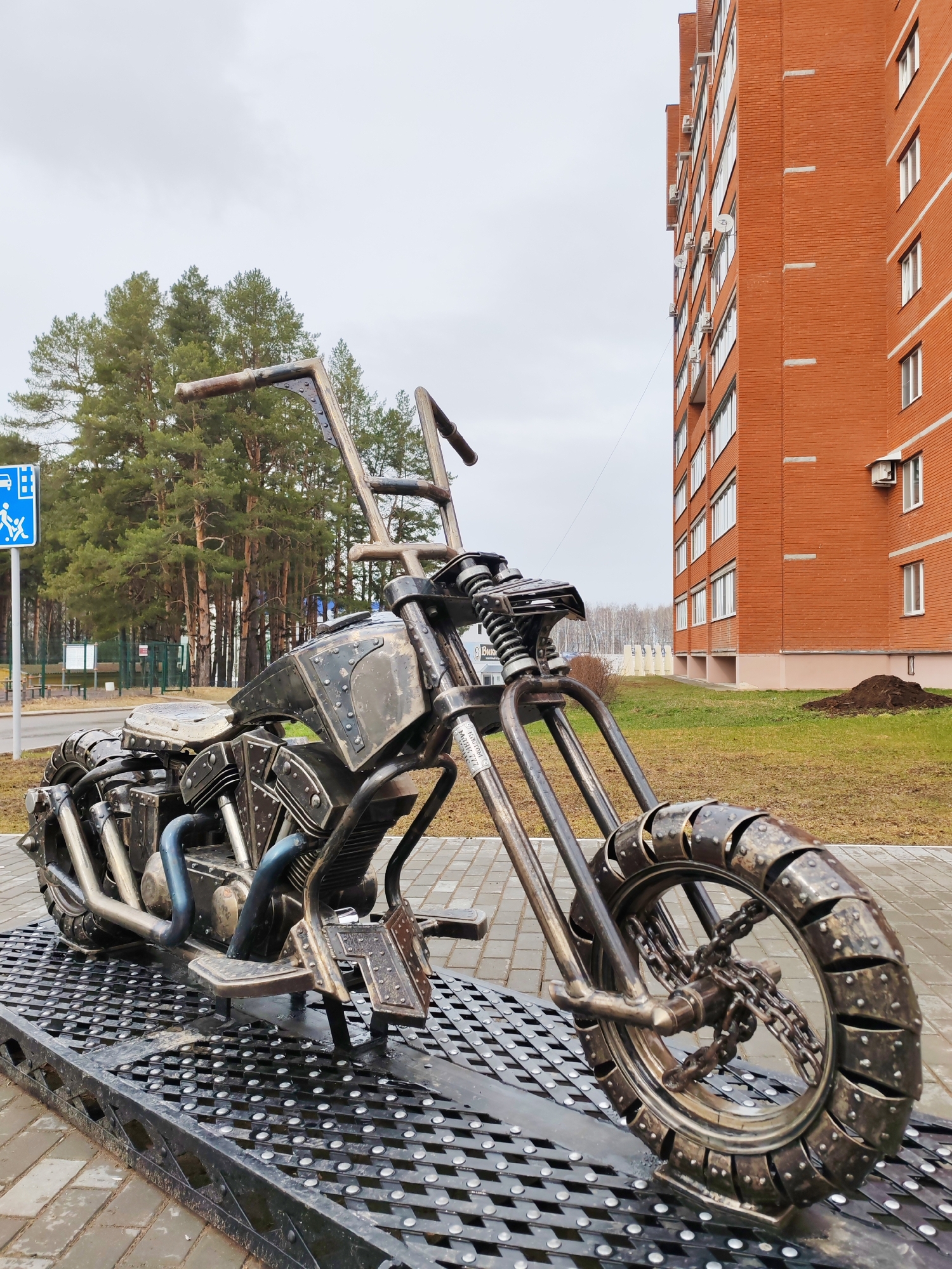 The image shows a metal sculpture of a motorcycle, likely a chopper-style bike, situated on a metal grid platform. The sculpture features details like a spring-fork front suspension, a V-twin engine representation, and high-rise handlebars. The wheels are constructed from metal components, including chain links.
In the background, there is a red brick apartment building. The sky is overcast. A blue and white sign indicating a pedestrian crossing can be seen next to a road. This image was taken in the city of Chaikovsky, Russia.