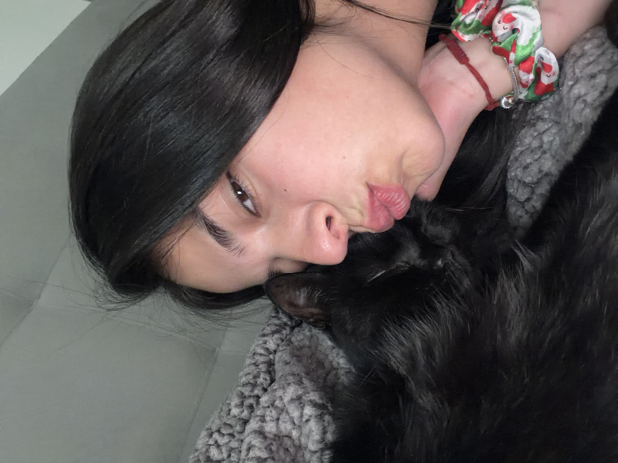 A woman with dark hair is lying on a gray, textured blanket with her face close to a black dog. She has a red bracelet and a scrunchie with a festive pattern on her wrist. Her lips are pursed. The location is not discernible as Gabrovo, Bulgaria.
