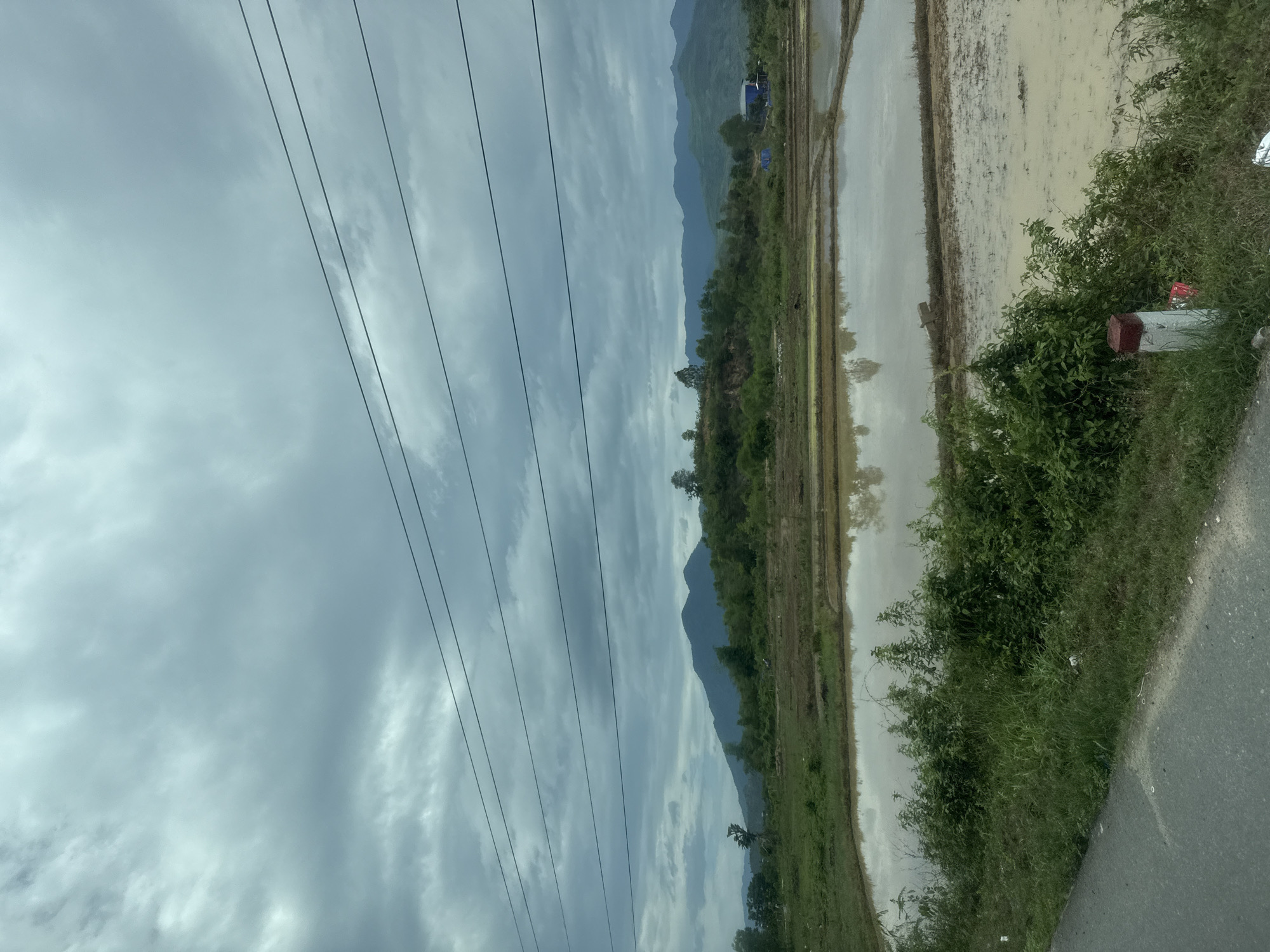 A landscape photograph captured in Diên Thọ, Vietnam, presents a serene rural vista under an overcast sky. The scene is dominated by natural elements, with man-made infrastructure also present.
In the foreground on the right, a dark asphalt road curves slightly, bordered by a strip of lush green grass and dense bushes. Partially obscured by the foliage, a white and red boundary marker or post is visible near the roadside. To the left of the road and vegetation, a wide expanse of flooded rice paddies stretches into the mid-ground, reflecting the cloudy sky above. The calm water surface shows reflections of distant trees and the muted grey sky.
Further into the mid-ground, a line of dense green trees and vegetation creates a natural division, behind which majestic, hazy mountains rise in the distance, their peaks blending into the soft, grey-blue atmosphere. Near the base of these mountains, a small blue tarp or structure can be faintly seen.
The sky above is largely covered by a thick layer of grey and white clouds, indicating an overcast day with diffused light. Several dark, parallel power lines traverse the upper portion of the image, running diagonally across the cloudy sky.
No people are visible in the image, and there are no specific activities taking place, creating a peaceful and still ambiance. The soft lighting suggests it is daytime, possibly late morning or afternoon, without direct sunlight. The landscape, with its rice paddies and mountains, strongly reflects the typical rural environment of Vietnam.