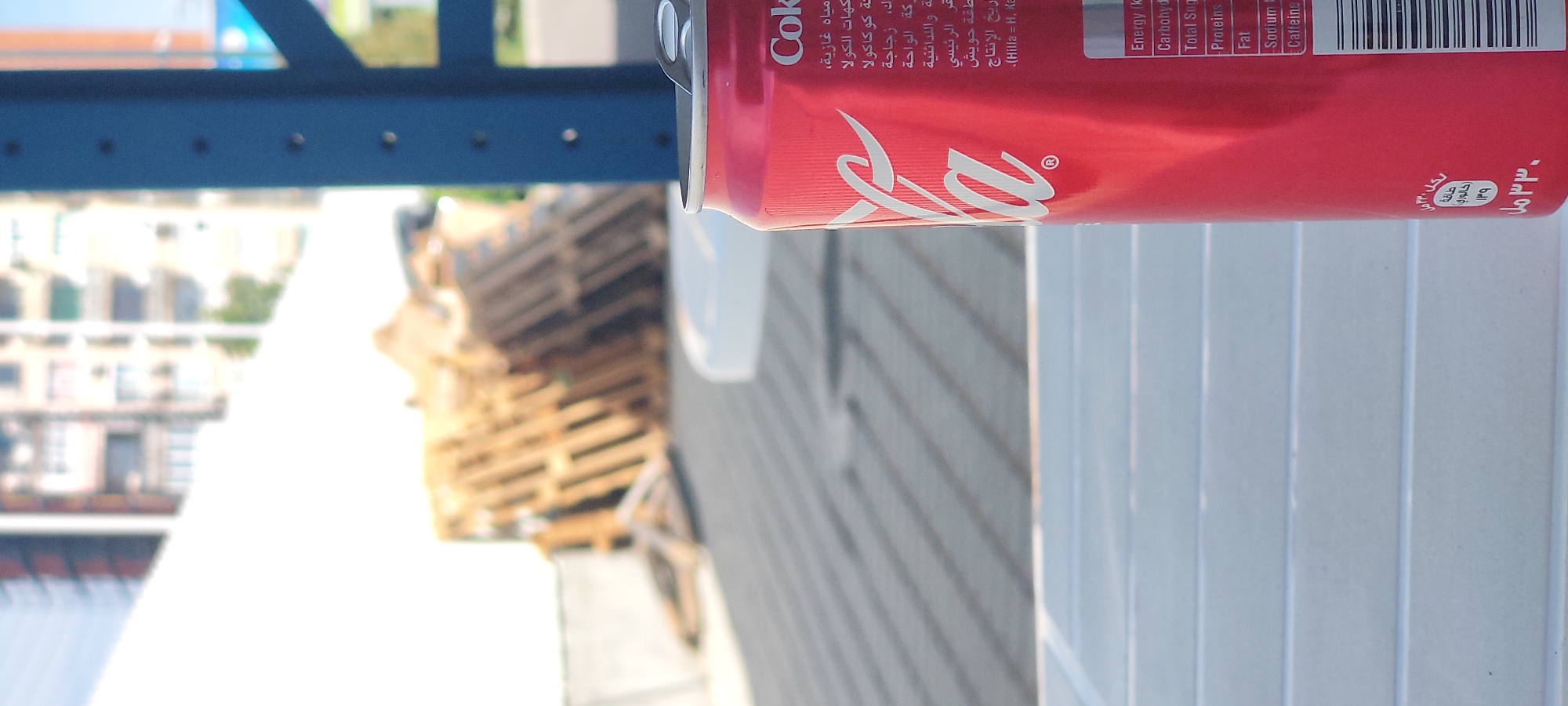 Here is a concise description of the image:
The image shows a red Coca-Cola can with Arabic writing on its label. The can is situated in what appears to be an urban setting, with blurred buildings and possibly some wooden pallets visible in the background. A blue structure is also partially visible at the top of the image. The setting might be near a construction or industrial area.