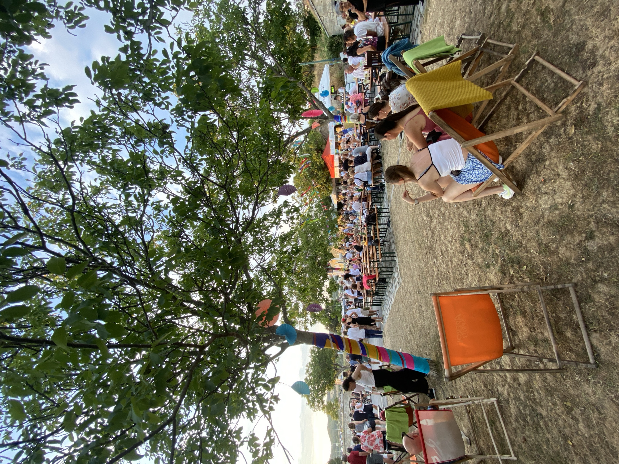The image depicts an outdoor gathering in Purbach, Austria, likely a festival or market, set during daytime under a tree canopy. The foreground features several empty and occupied lounge chairs made of wood and fabric. Two women are seated in lounge chairs in the middle ground, facing away from the camera, with one woman wearing a white tank top and blue and white patterned skirt, and the other in a pink tank top. A yellow blanket is draped over one of the chairs, and a green blanket over another.
In the background, a large crowd of people are seated at picnic tables, suggesting a dining or social event. There are also various tents and stalls visible, some with colorful decorations. A tree in the left foreground is wrapped with colorful fabric and adorned with hanging decorations, including what appear to be paper umbrellas. The overall atmosphere suggests a relaxed, community-oriented event. The ground is covered in dry grass and dirt, and trees with green foliage form a significant part of the environment. The sky is partly cloudy.