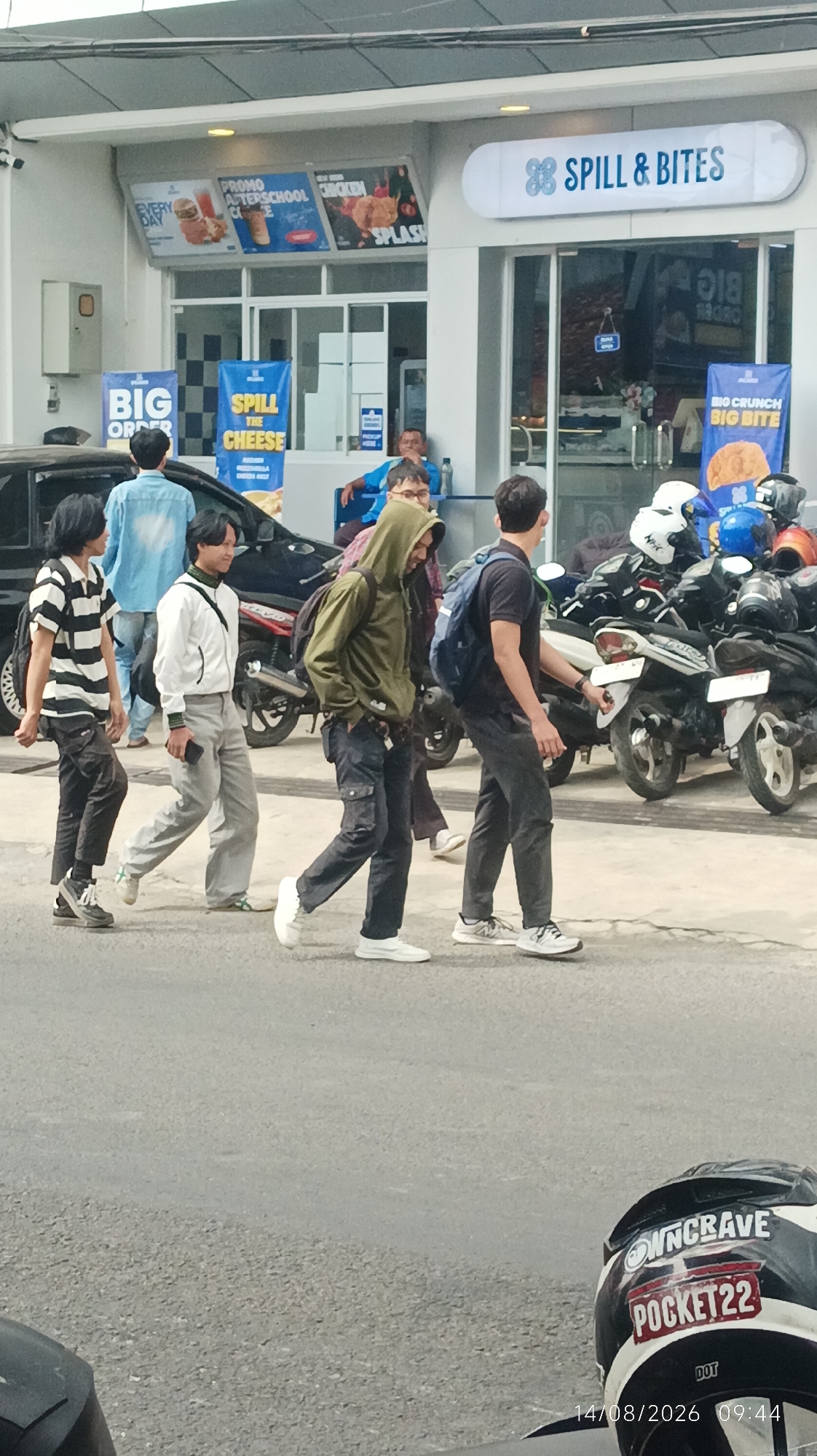 A group of young men walks by a storefront that reads "SPILL & BITES". The men are casually dressed, with some wearing hoodies and others wearing t-shirts. One man is wearing a white jacket and gray pants. The storefront appears to be a fast-food restaurant, with posters advertising food items like chicken and burgers. In the background, there are several motorcycles parked, suggesting a busy urban environment. The ground is paved, and there are signs indicating "BIG ORDER" and "SPILL THE CHEESE" outside the establishment. The overall impression is a snapshot of everyday life in a bustling Indonesian city. The timestamp on the bottom of the image indicates the date and time as 14/08/2026 09:44.
