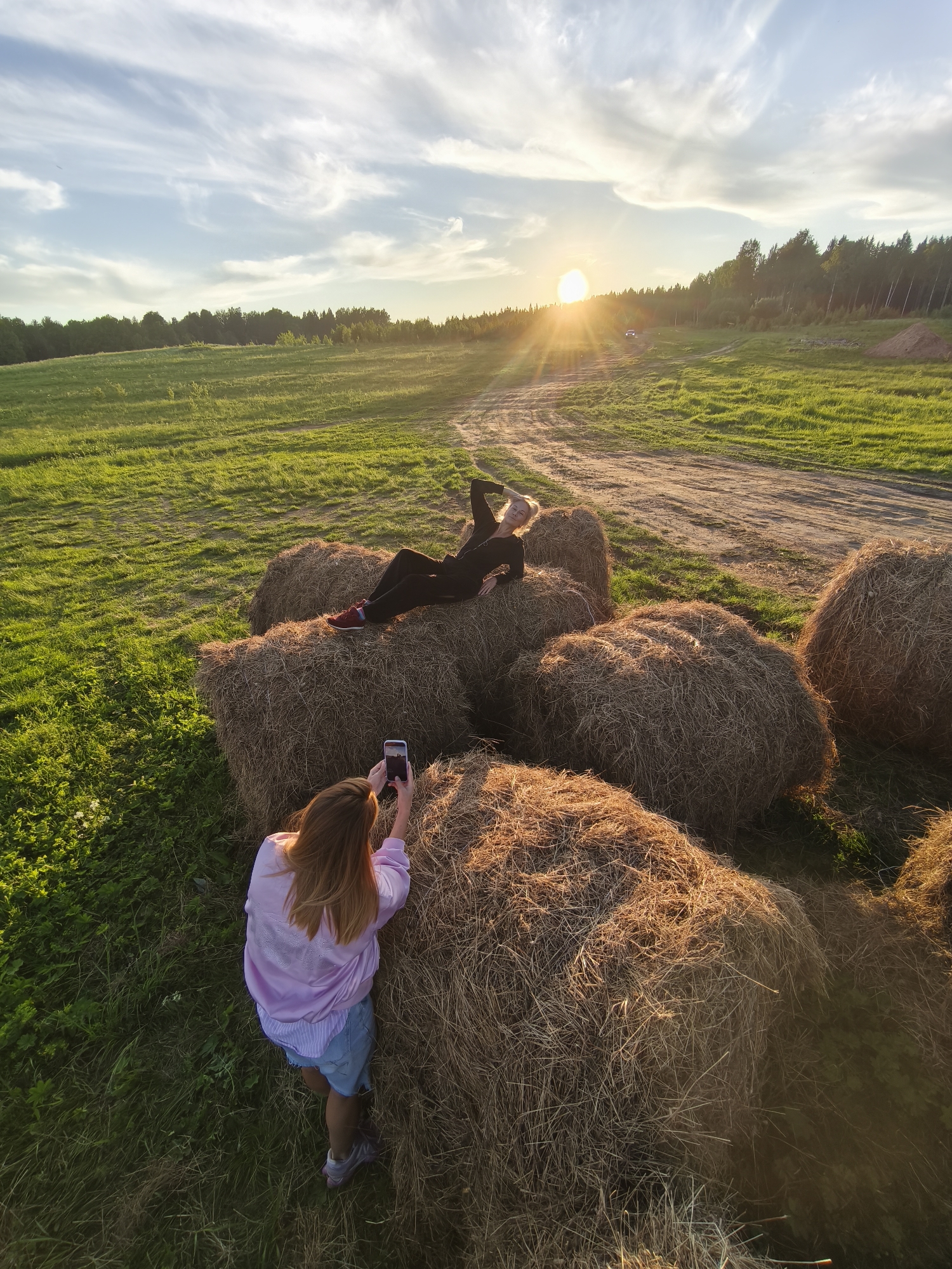 This outdoor photograph captures two women amidst a rural landscape during sunset in what appears to be Стеклянный, Russia. One woman with blonde hair, dressed in dark clothing, is relaxed and posing, lying on a cluster of large, round hay bales. Her arm supports her head as she looks towards the horizon. The second woman, seen from a high angle and from behind, has long brown hair and is wearing a light purple jacket over a striped top and shorts. She is actively taking a photo of the first woman with a white smartphone.
The scene is set in a wide, lush green field with a winding dirt path. A dense forest line stretches across the distant horizon. The sky is a mix of pale blue and white, adorned with wispy clouds. The most striking notable detail is the golden hour light of a setting sun, which is prominently visible on the horizon, casting a warm glow and long shadows across the field and hay bales. A bright lens flare emanates from the sun. A small white car is visible in the distance on the dirt path. The overall impression is one of leisure and enjoyment of the natural environment during a beautiful evening. No discernible text is visible in the image.