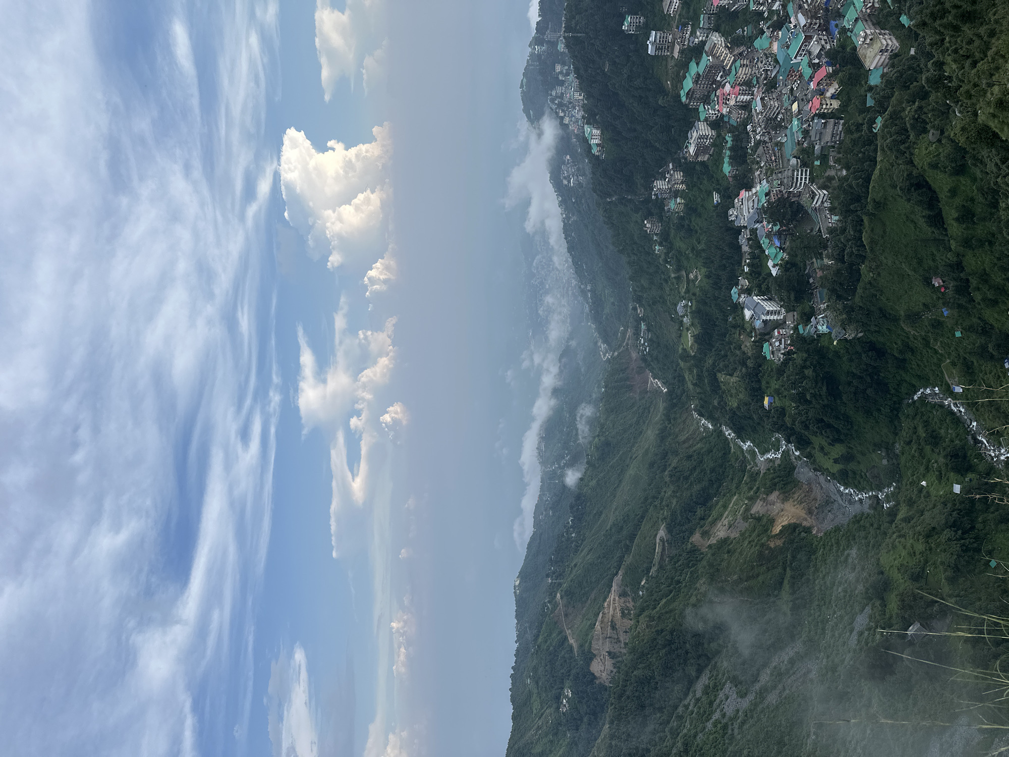 The image is a high-angle, wide-shot, landscape view of Bhagsu, India, showcasing a valley with a winding river and a town built into the hills. The sky above is a gradient of blue, with scattered white clouds, some of which appear wispy and others more cumulus-like.
In the foreground, the lush green slopes of the mountains dominate. A thin, rocky cliff face is visible on the left, with evidence of human alteration, possibly for construction or road building. A small river snakes its way through the valley, appearing as a white ribbon against the dark green foliage and rocky terrain.
Further into the valley, the town of Bhagsu begins to appear. Buildings with colorful roofs, predominantly green, red, and grey, are nestled into the steep hillsides. The architecture suggests a mix of residential and commercial structures, with multiple stories and balconies visible. The town extends across the valley and up the slopes, demonstrating its adaptation to the mountainous topography.
In the mid-ground and background, the valley continues, with more buildings and development visible. Patches of mist or fog are present, obscuring some of the distant landscape and adding a sense of depth and atmospheric haze. The overall environment is a blend of natural mountainous terrain and human settlement. No people are clearly discernible in the image.