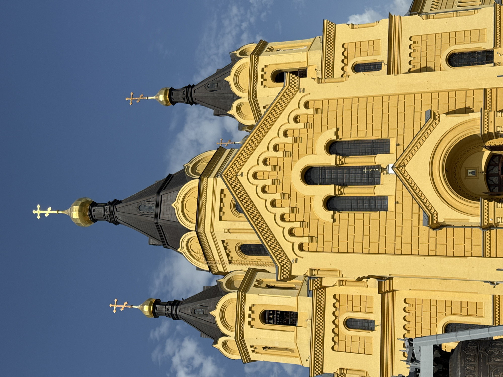 The image shows the Alexander Nevsky Cathedral in Nizhny Novgorod, Russia. The photograph is oriented vertically. The cathedral is painted a yellow color, with three black onion domes topped with golden crosses. The sky is blue and partly cloudy. The building is highly decorated with repeated geometric patterns and arched windows. The building is large, and a piece of machinery is located in the lower-right corner.